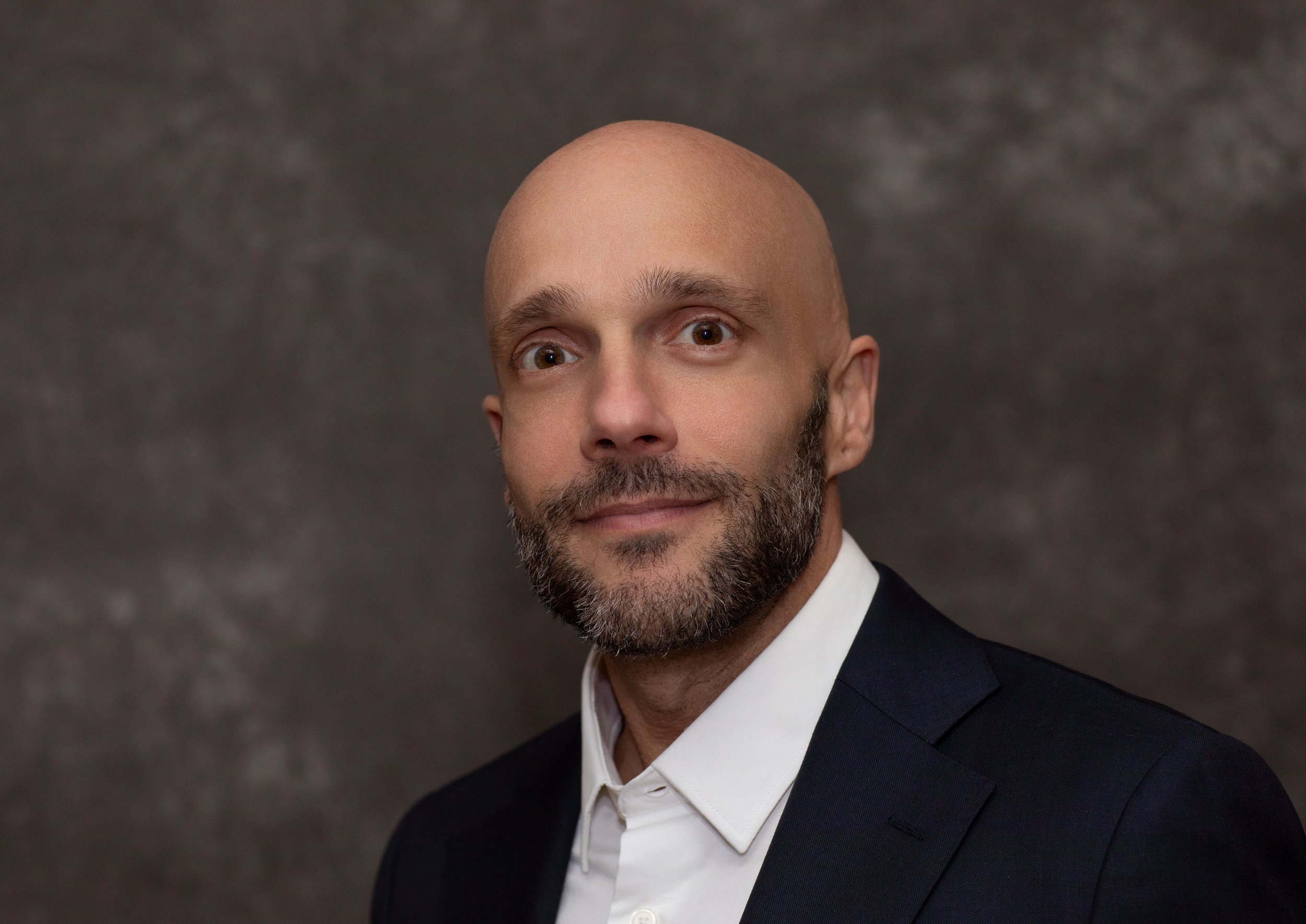 A bald man with a beard in a suit and white shirt on a dark textured background.