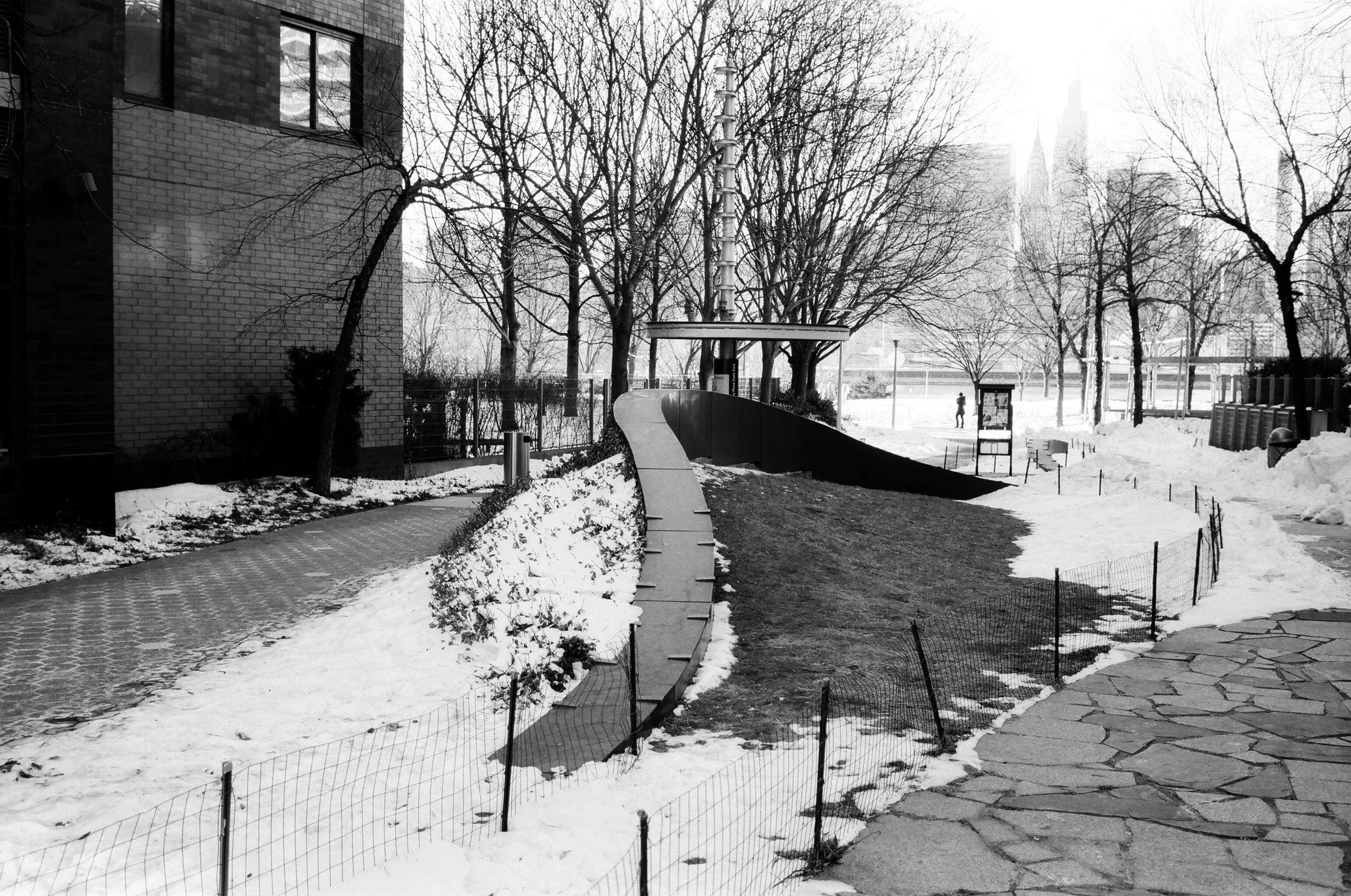 A snow-covered urban park with a winding pathway, leafless trees, and a skateboard ramp in the center. Buildings are visible in the background.