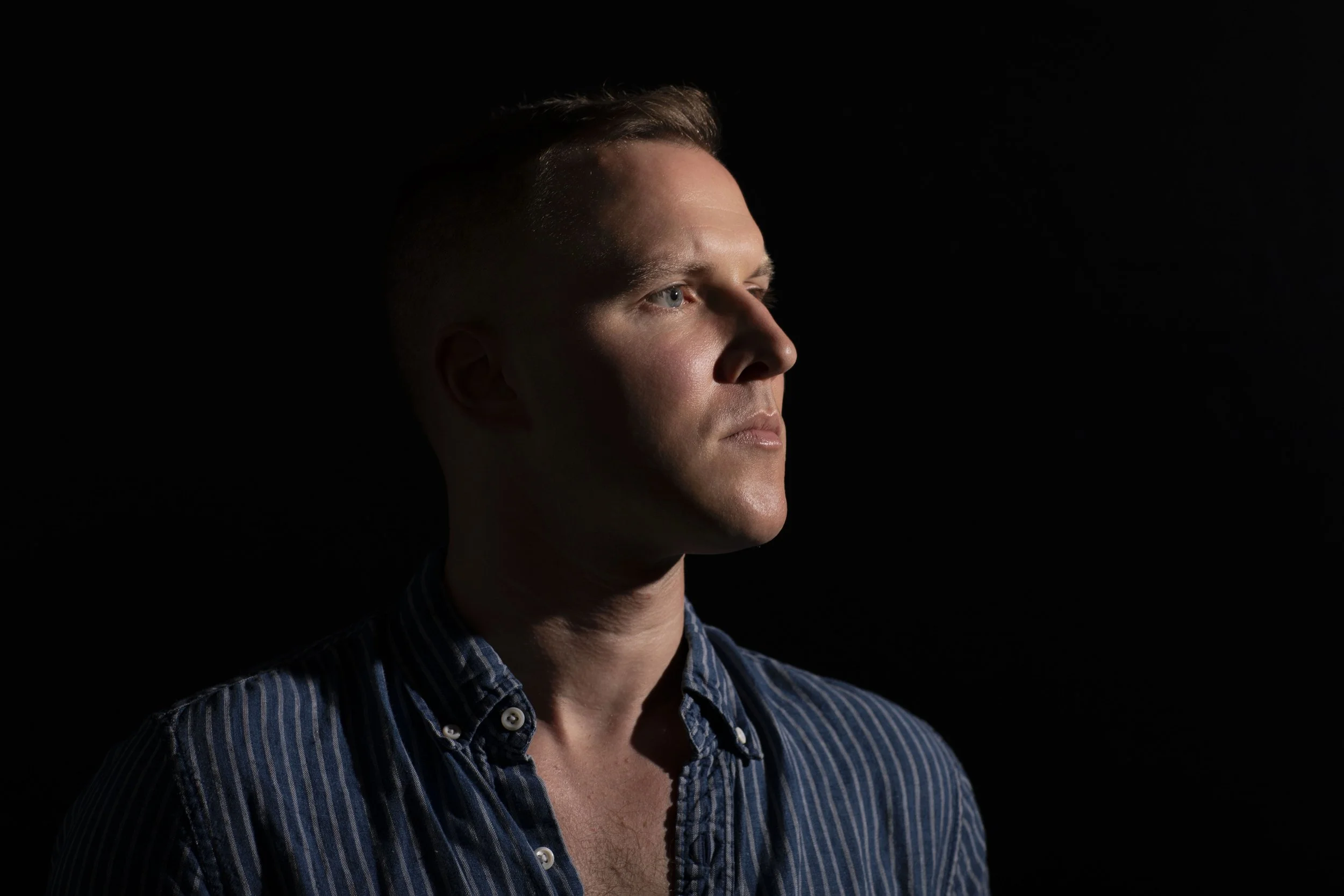 Portrait of a man with light skin and short brown hair, wearing a blue and white striped shirt, facing right in low-light setting.