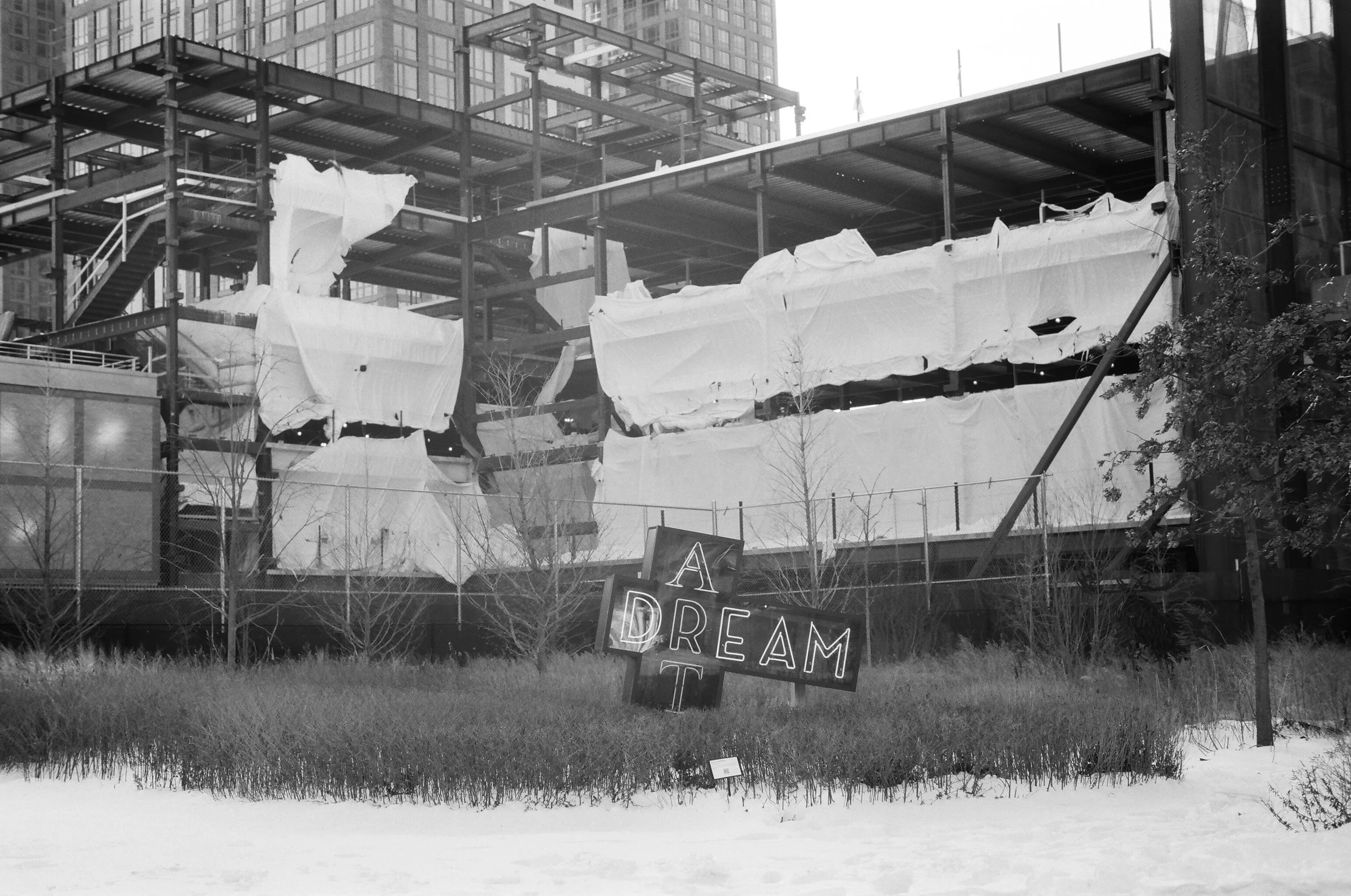 A construction site with a scaffolding structure covered partially by white tarps, surrounded by a chain-link fence. In front, a sign reads 'A DREAM T' with some letters obscured, in front of a snow-covered area with small bushes and leafless trees.