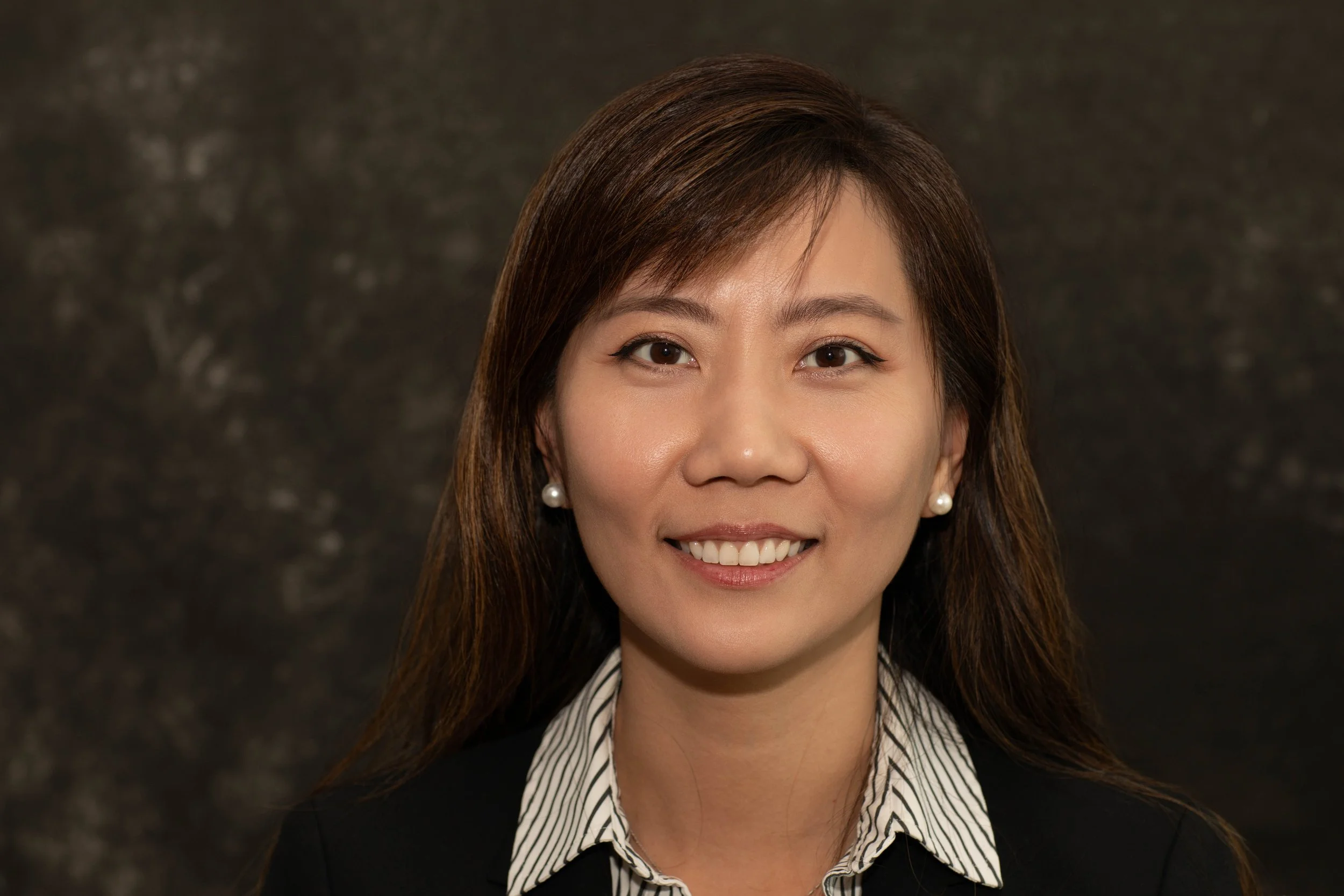 Portrait of an Asian woman with shoulder-length brown hair, wearing pearl earrings and a black blazer over a striped collared shirt, smiling in front of a dark textured background.