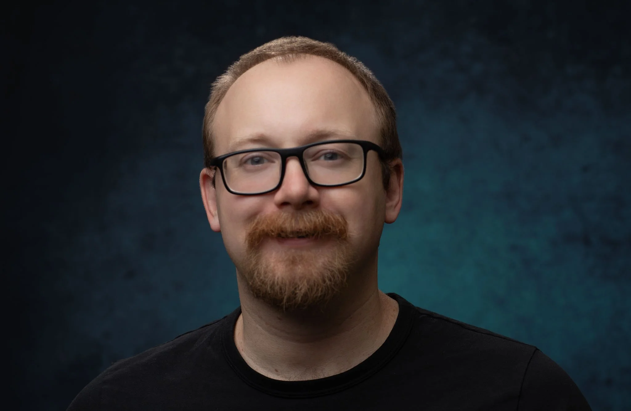 A portrait of a young man with glasses and a beard, smiling, wearing a black t-shirt, against a dark textured background.