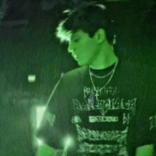 A young man with dark hair wearing headphones and layered necklaces, illuminated in green lighting.