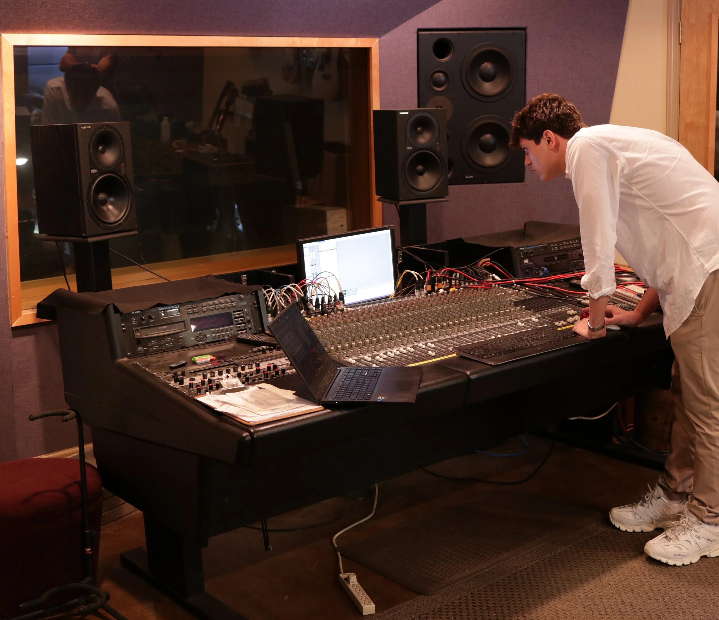 A young man is working at a large audio mixing console in a recording studio with several speakers around him, a laptop, and a computer monitor displaying audio software.