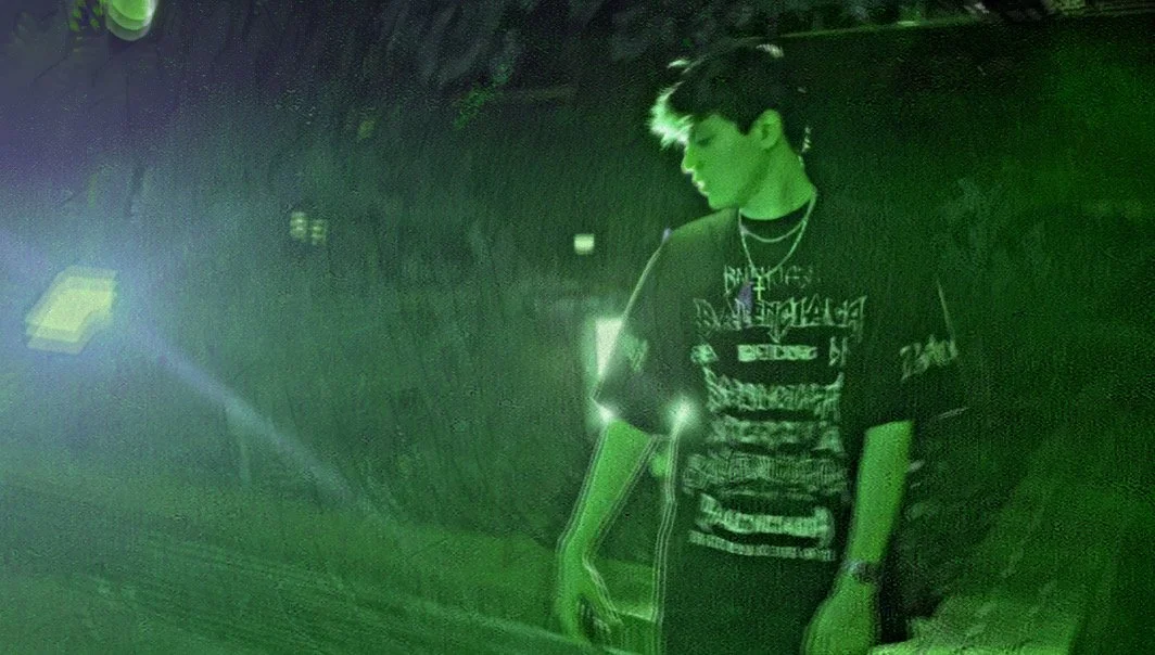 A young person standing outdoors at night illuminated by green light, wearing a graphic T-shirt and a chain necklace, with dark, wavy hair.