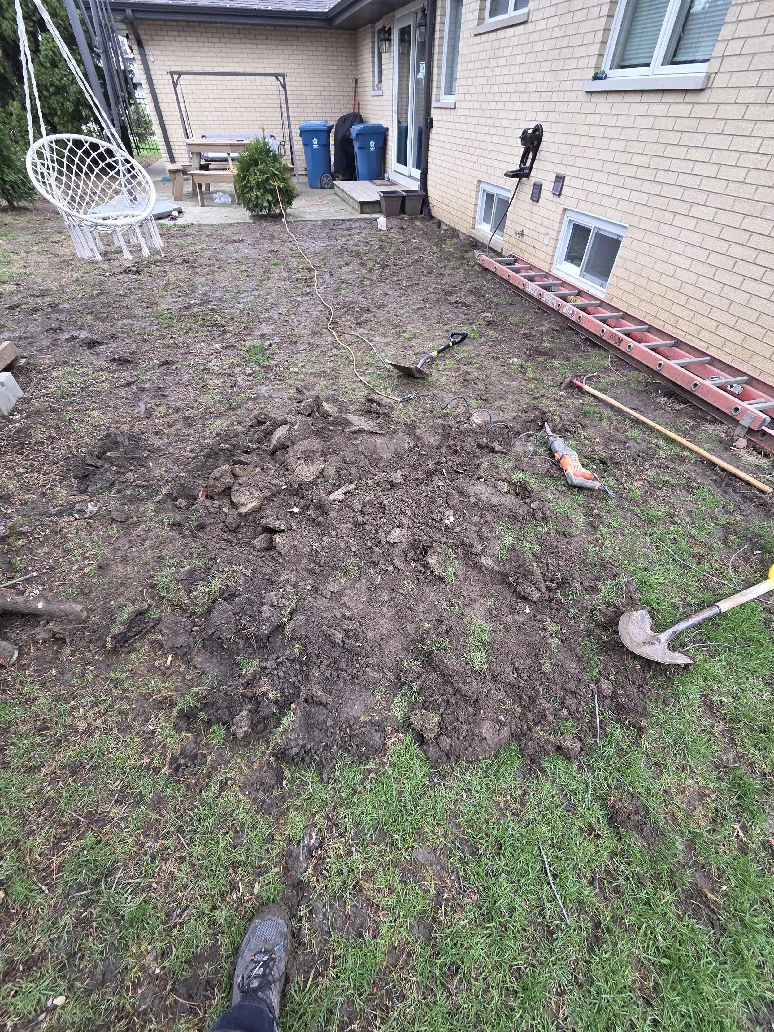 Backyard with dirt and grass, tools, ladder, and outdoor furniture during yard work.