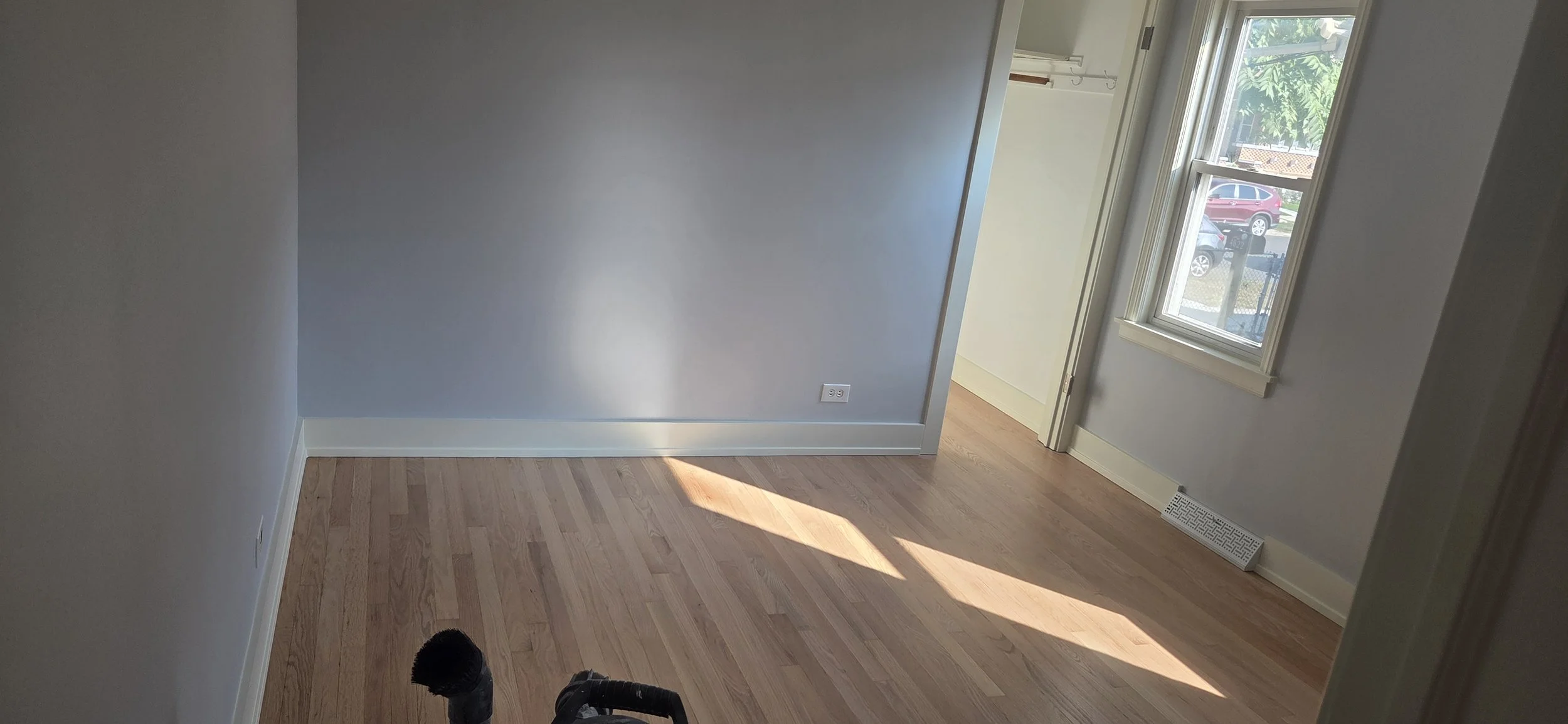 Empty room with hardwood floors, white walls, a window showing parked cars outside, and sunlight coming through the window.