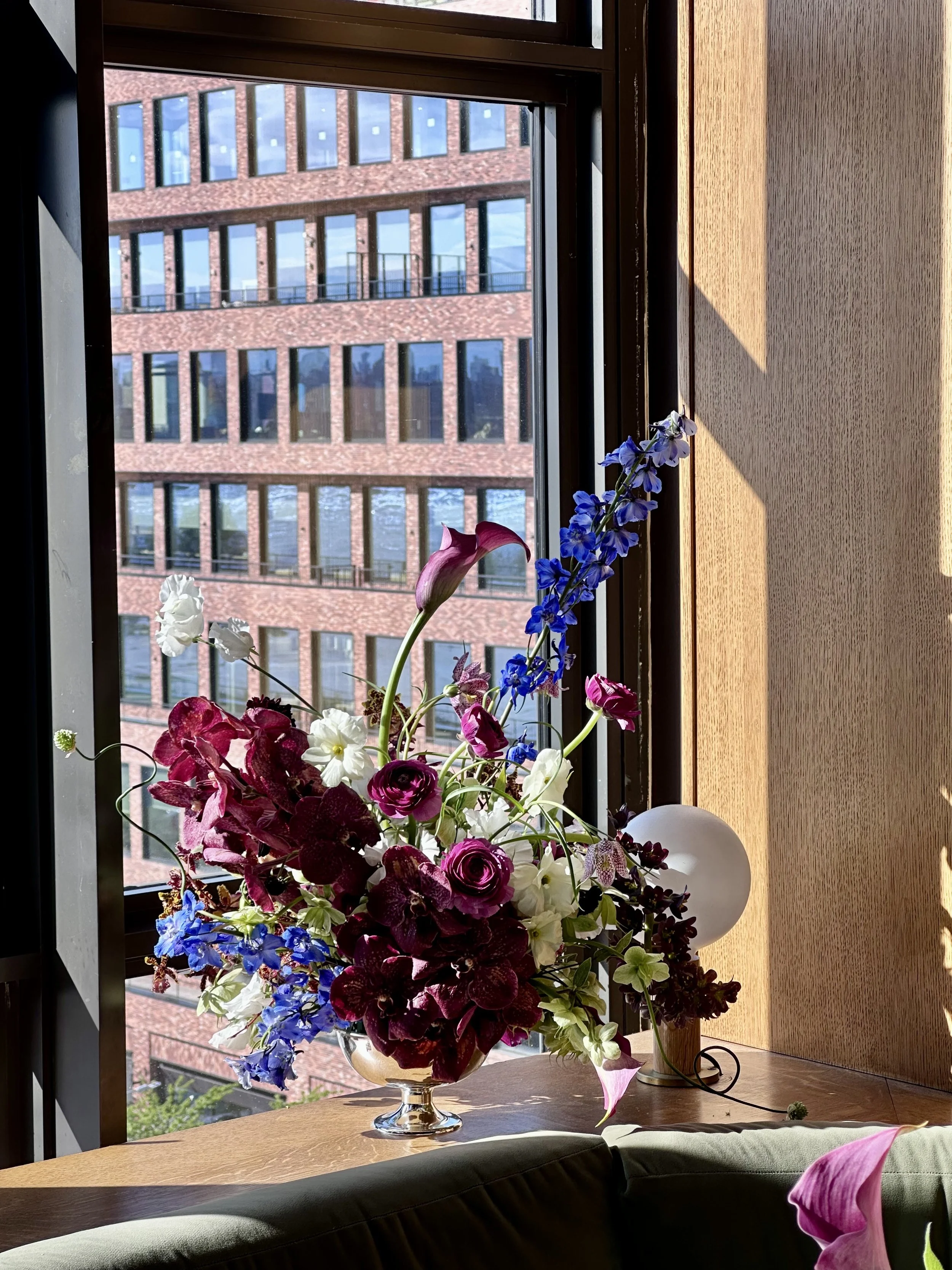 Various vases with colorful flowers, including pink, yellow, and white roses, displayed on a table and stand outdoors with green bushes and a wooden fence in the background.