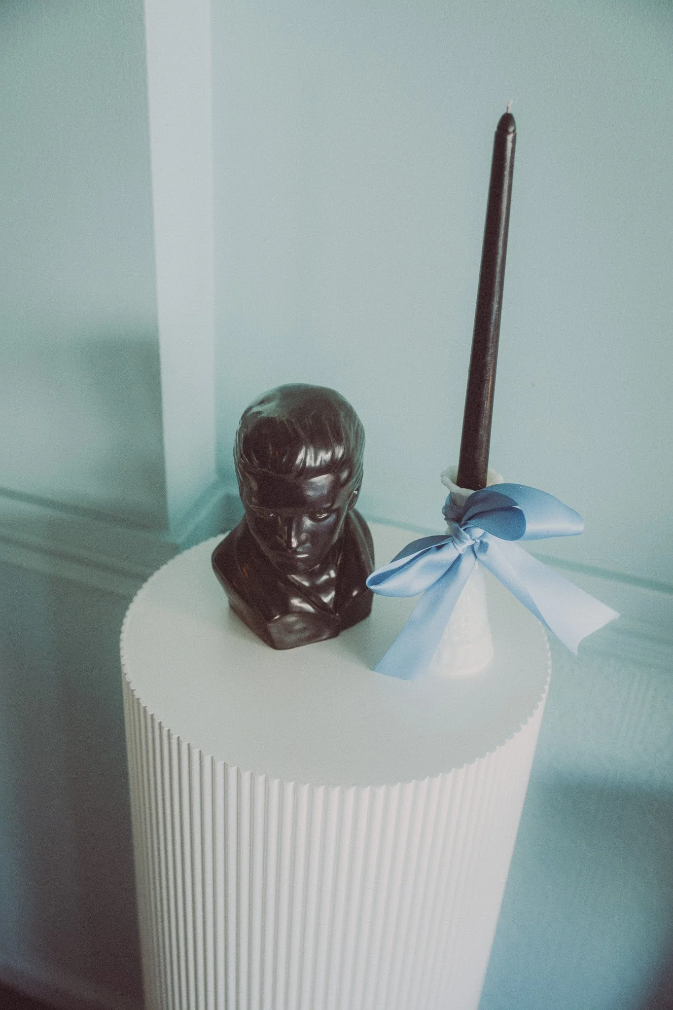 A bust sculpture of a person's head and shoulders made of dark material, and a tall dark candle in a white holder with a light blue ribbon tied around it, are placed on a white cylindrical surface.