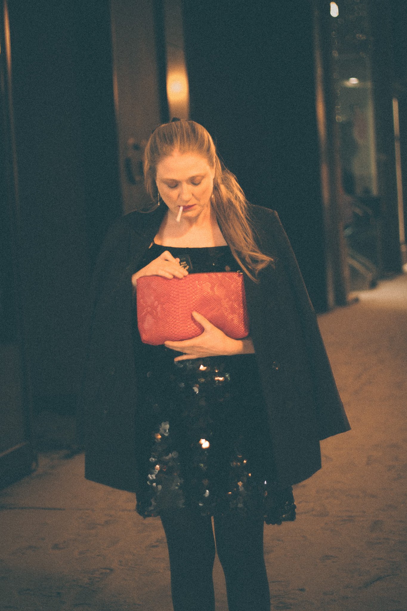 A woman with long hair, dressed in black, is holding a red clutch and smoking a cigarette in a dimly lit indoor setting.