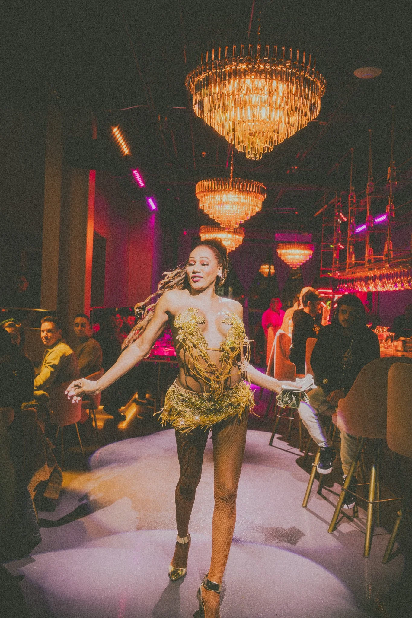 A woman in a revealing gold dress is dancing on a stage in a nightclub with chandeliers and colorful lighting.