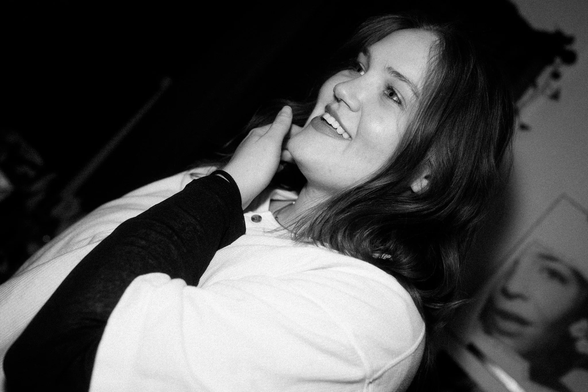 A black and white photo of a smiling woman with shoulder-length hair, wearing a light shirt and a dark long-sleeved shirt underneath, touching her face with her hand.