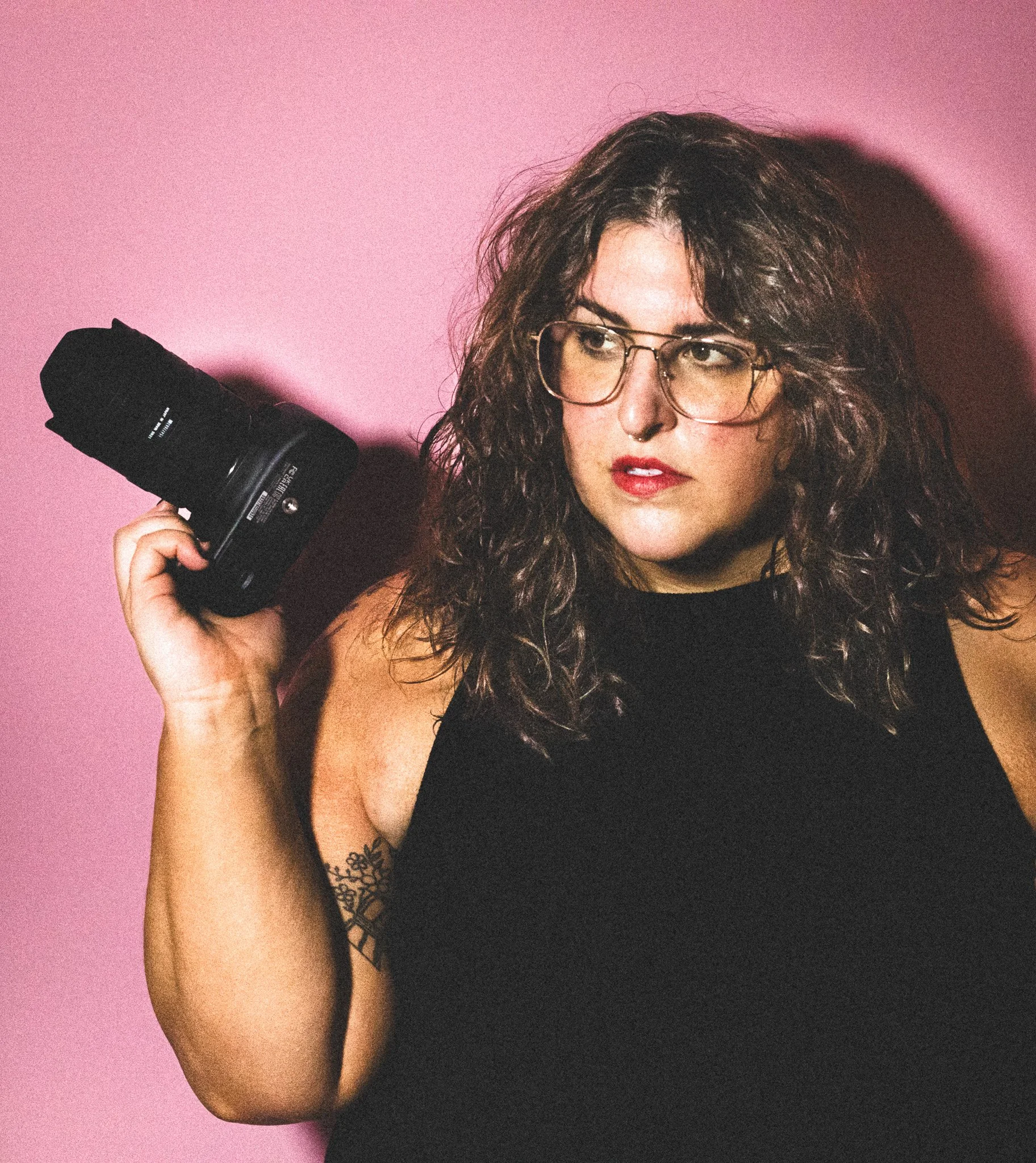 A woman with curly hair, glasses, and a septum piercing holding a camera against a pink background.
