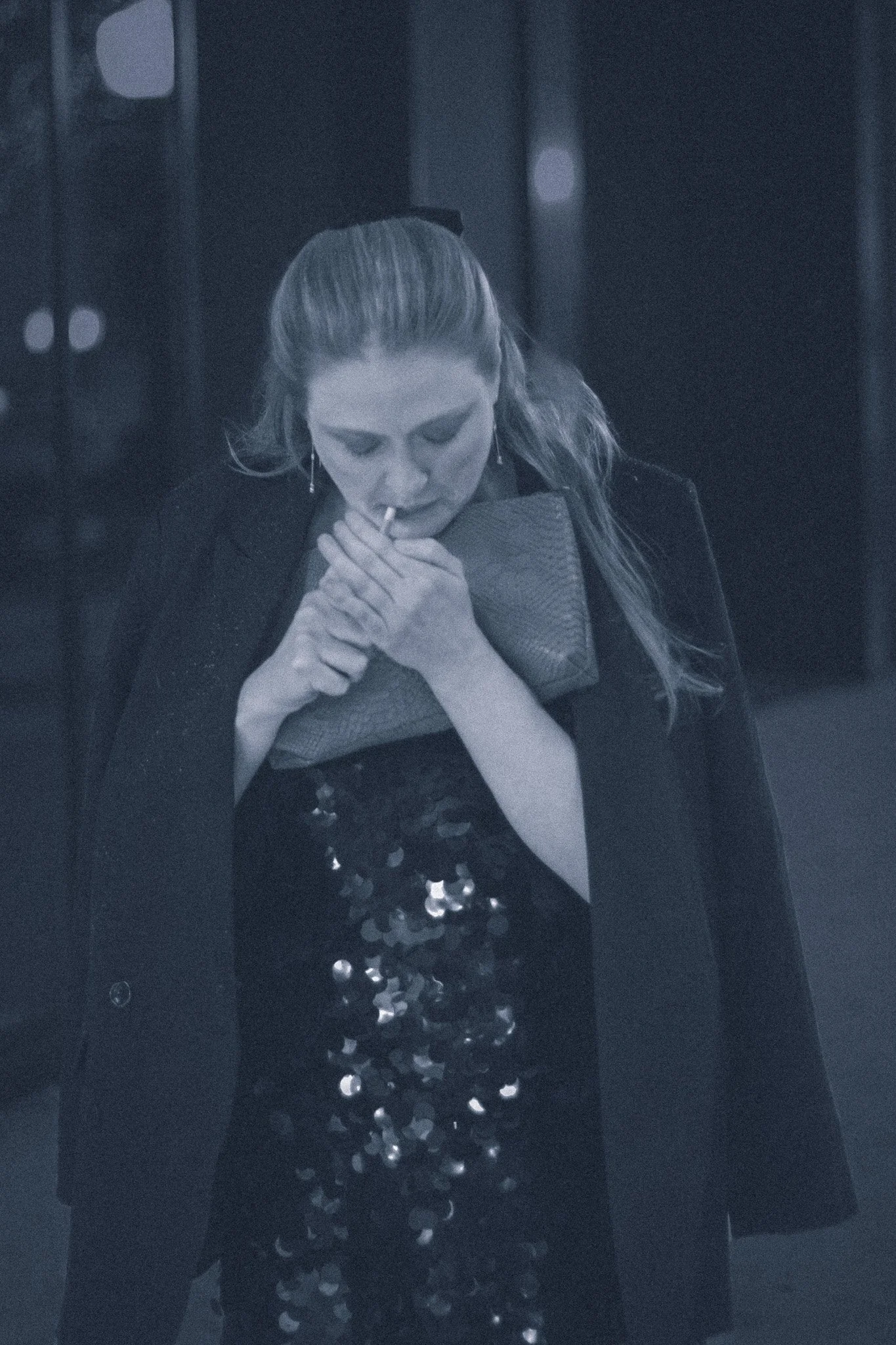 A woman with long hair, wearing a black dress with sequins, leaning on a clutch bag while lighting a cigarette with a matchstick in a dimly lit setting.