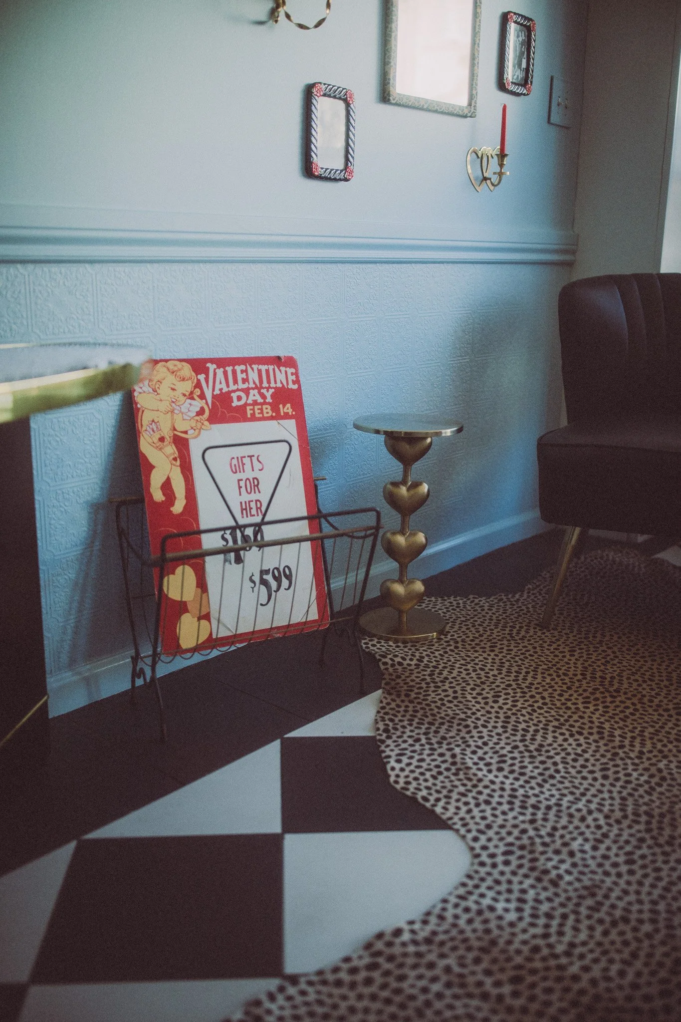 Valentine's Day promotional sign inside a vintage-style room with black and white checkered floor, leopard print rug, and eclectic decor, including framed mirrors and a gold heart-shaped candlestick.