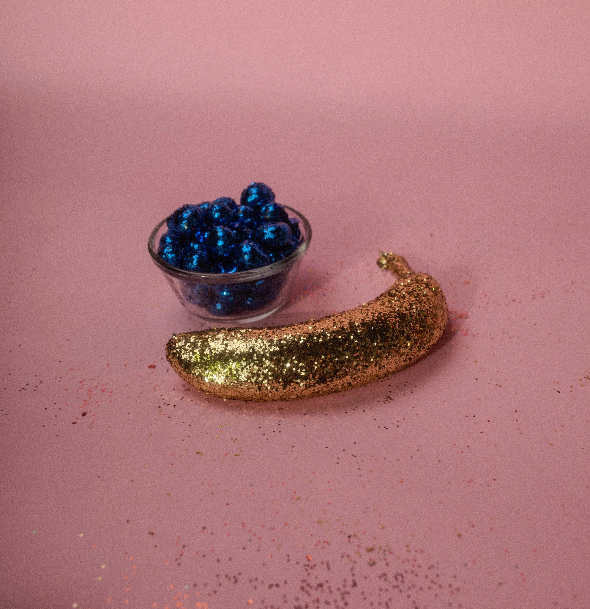 Decorative gold-glittery banana and a bowl of shiny blue beads on a pink background with scattered glitter.
