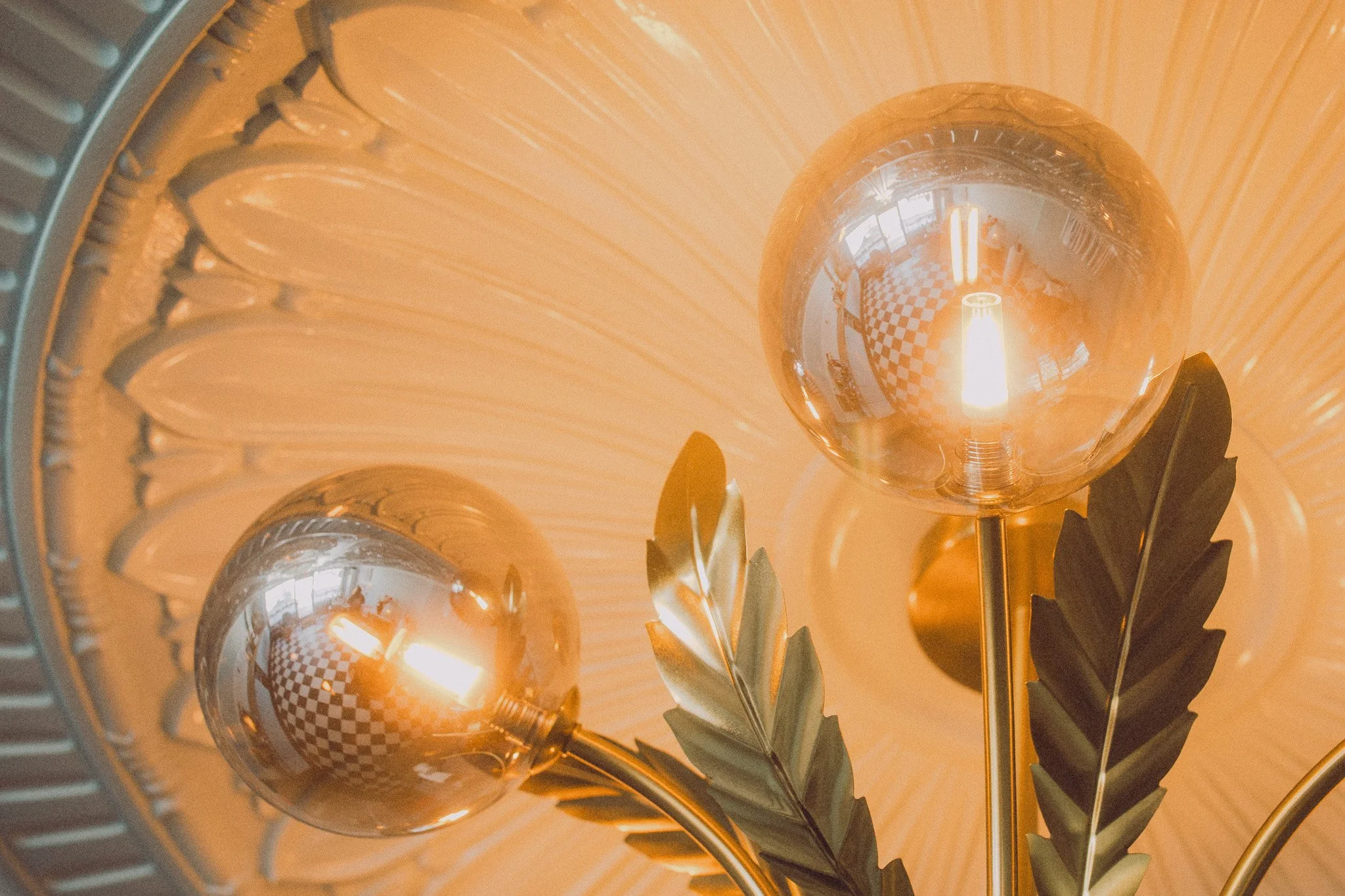 Close-up of a chandelier with spherical bulbs and decorative metal leaves, reflecting a checkered floor and a window.
