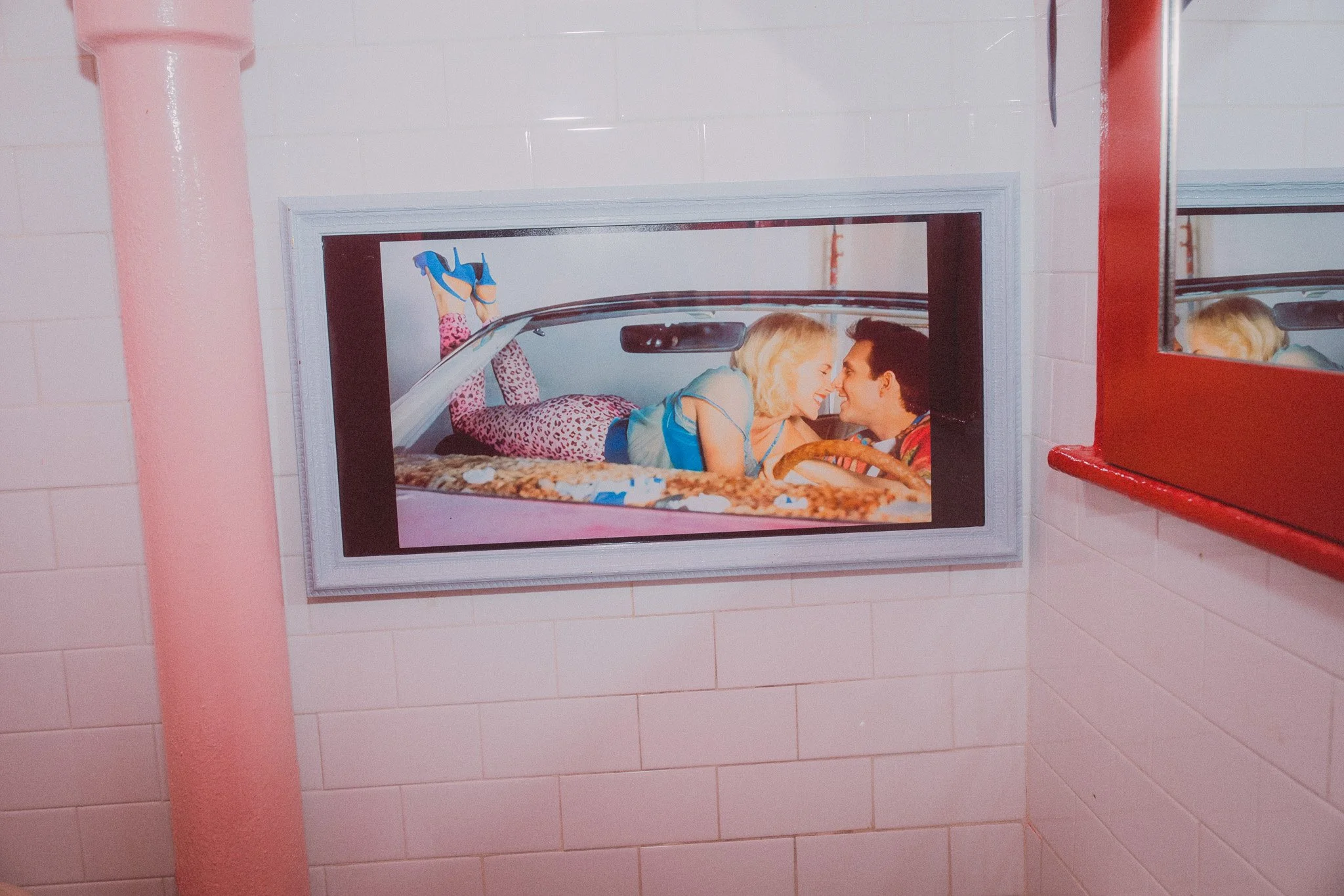 A framed picture on a pink wall shows a woman and a man smiling and leaning close to each other inside a car. The woman is wearing a blue top, and the man is wearing a multicolored shirt. The woman is lying on her stomach with her legs bent at the kn