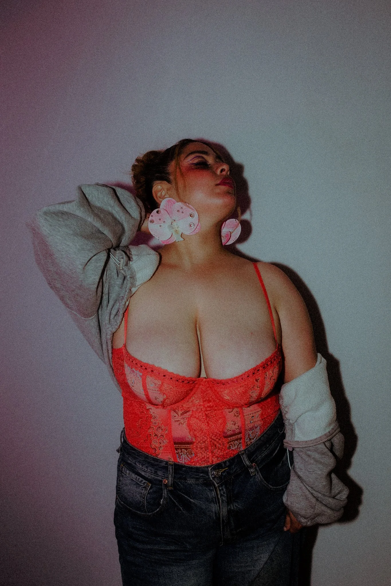A woman with dark hair and makeup posing against a plain wall, wearing a red lace bustier top, dark jeans, and a gray jacket draped over her shoulders, with pink butterfly earrings.