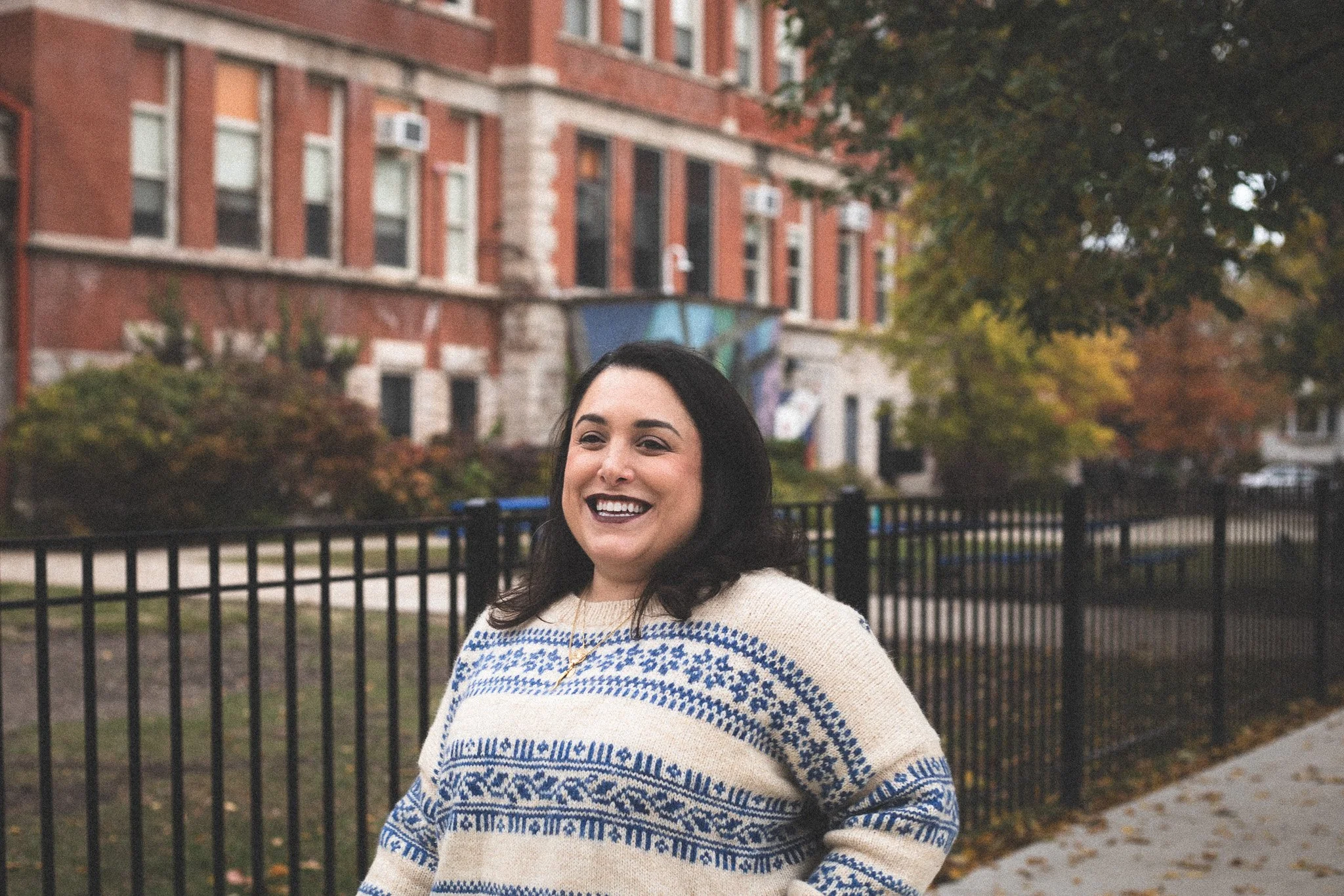 A woman with dark hair smiling and wearing a cream and blue patterned sweater, standing outdoors next to a black metal fence, with a brick building and trees with autumn foliage in the background.