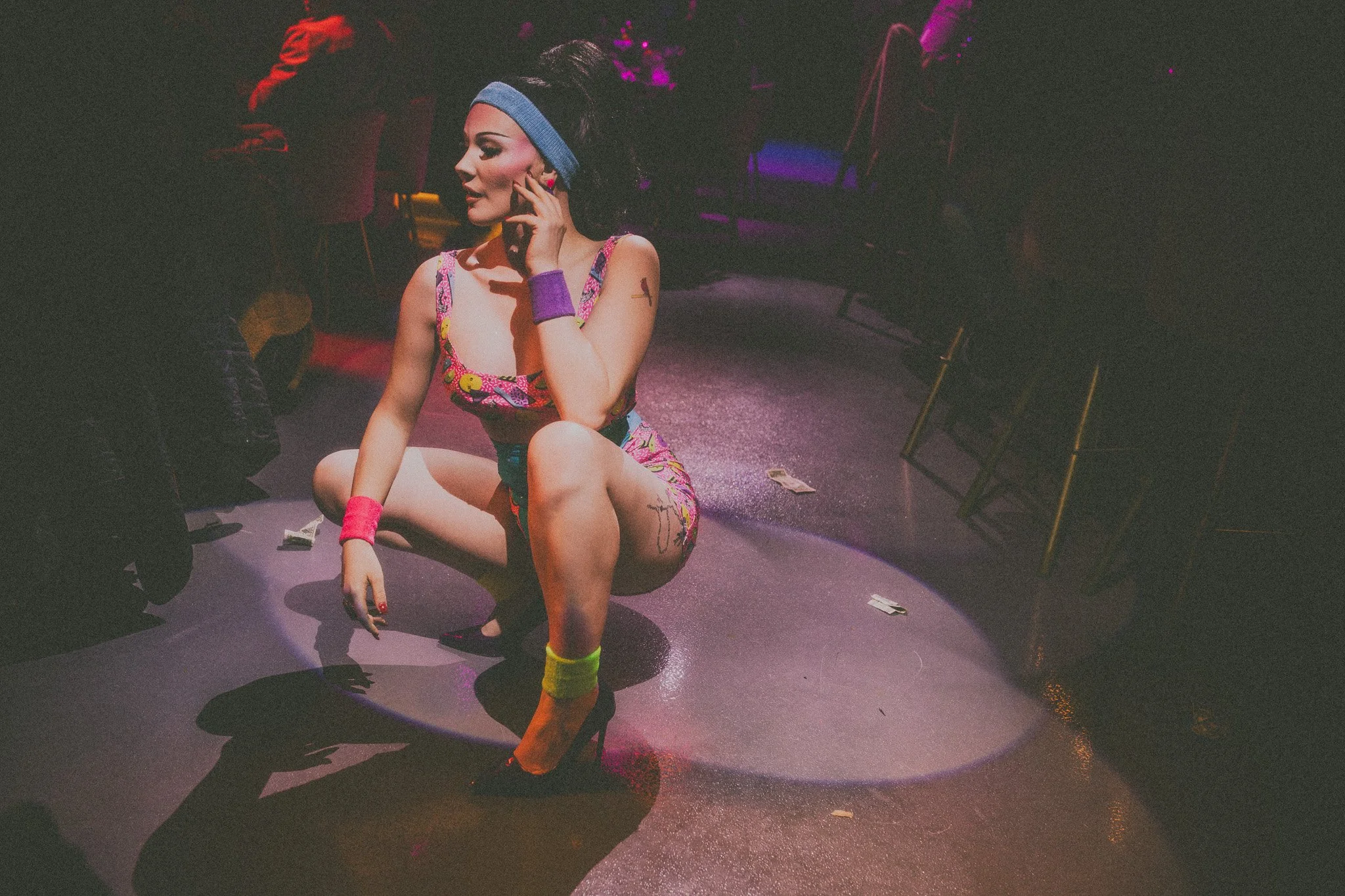 A woman in colorful, retro-style dance attire with neon accessories is on her knees on a stage, talking on a cellphone, with scattered dollar bills around her. The scene is dimly lit with purple and pink stage lights and chairs in the background.