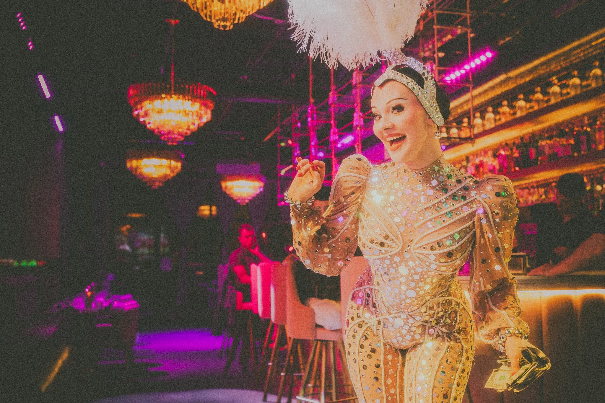 A performer in a shiny, decorated gold outfit with a feathered headpiece, smiling and gesturing joyfully in a colorful, neon-lit bar or nightclub.