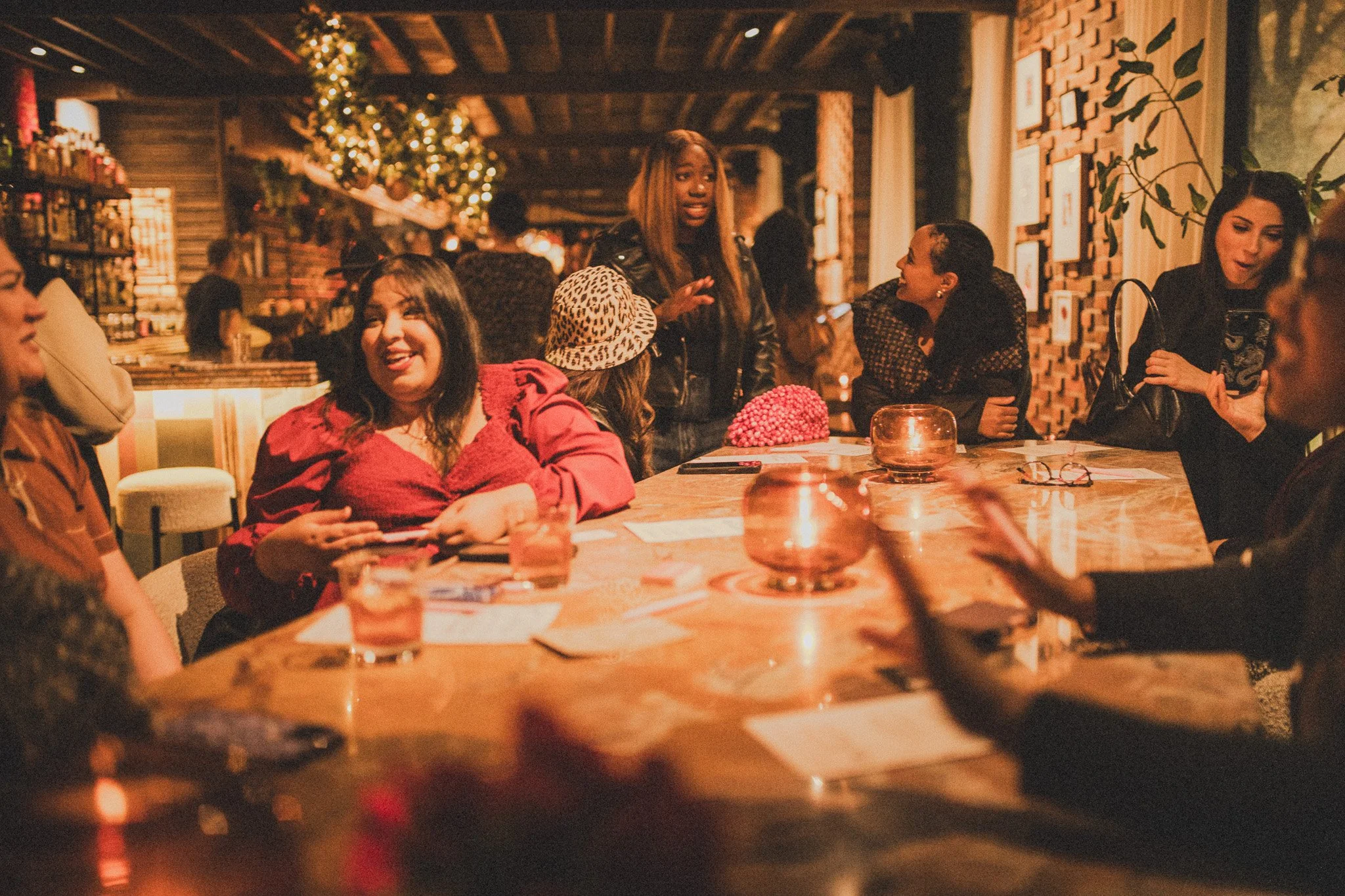 A group of women socializing around a table in a cozy, warmly lit restaurant decorated with festive holiday ornaments, including a Christmas tree with lights in the background.