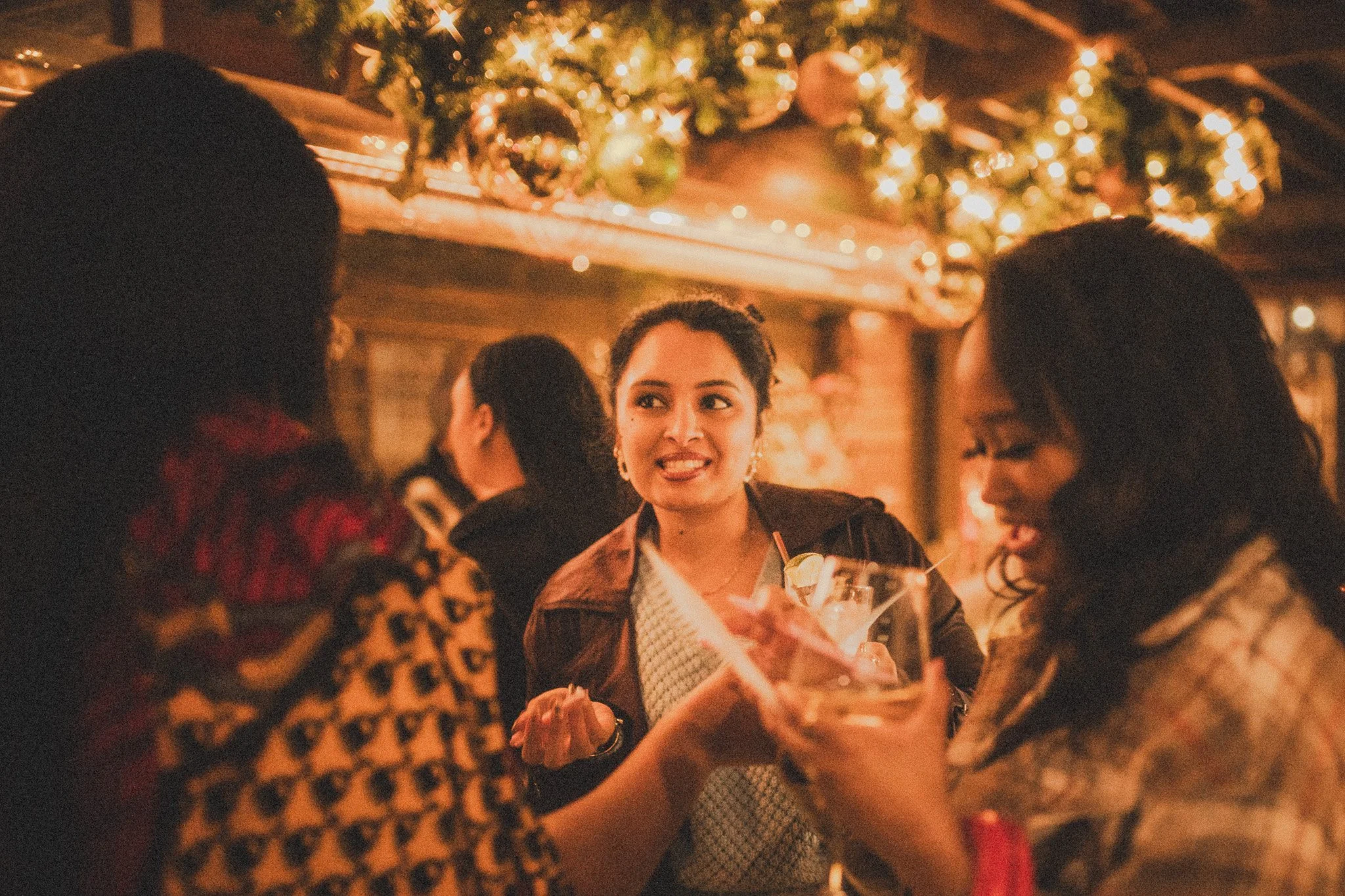 People socializing and holding drinks at a cozy, festive setting decorated with string lights and greenery.