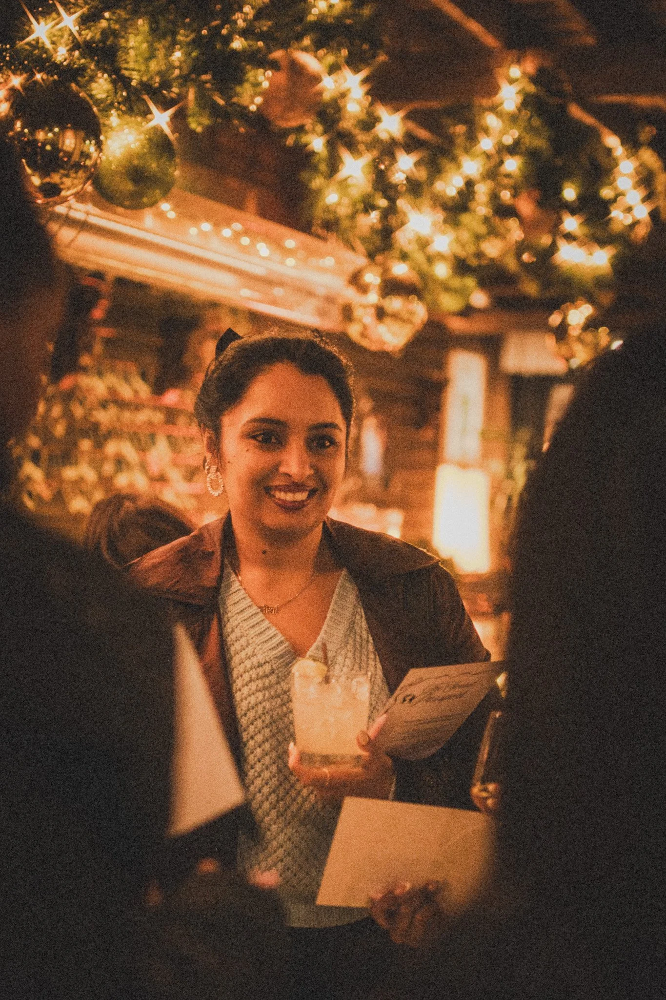 A woman smiling and holding a drink in a cozy, decorated Christmas setting with twinkling lights and ornaments.