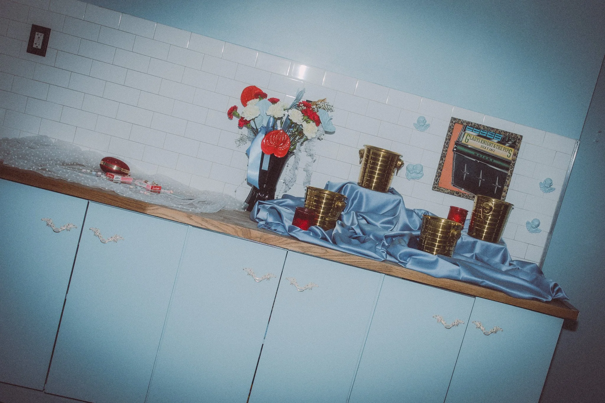 Decorative display on a kitchen counter with a flower bouquet, gold buckets, red candles, and a blue satin cloth, against a tiled wall.