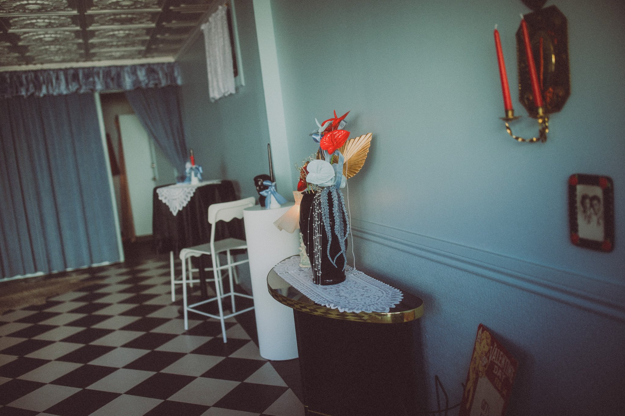 Interior of a room with checkered black and white floor, a small black table with a lace cloth, and a vase with artificial flowers, gold paper fan, and beads. Blue curtains on a window and a dark curtain covering part of the background. Wall-mounted 