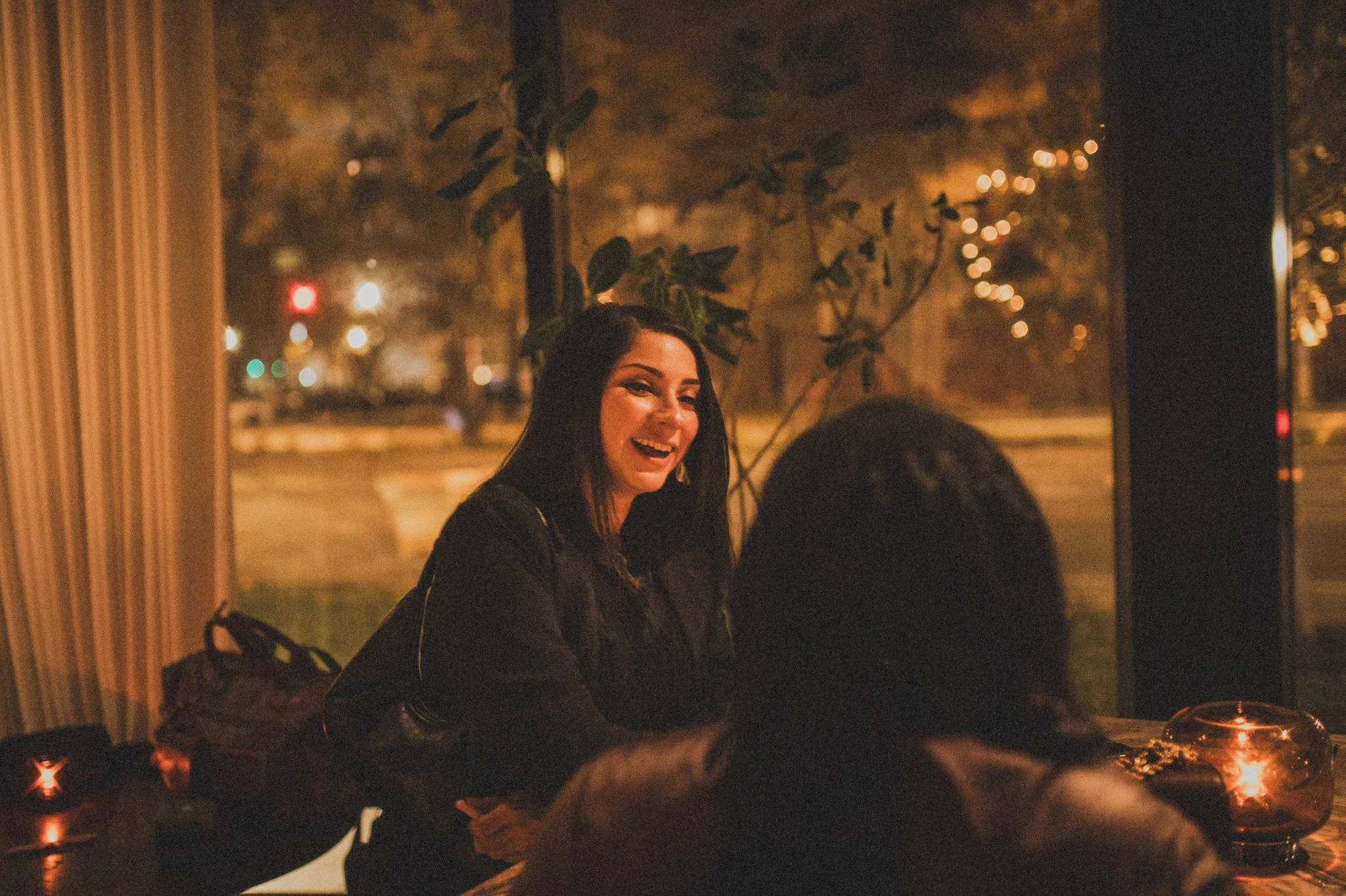 A woman with long dark hair smiling and talking to another person inside a dimly lit restaurant at night.