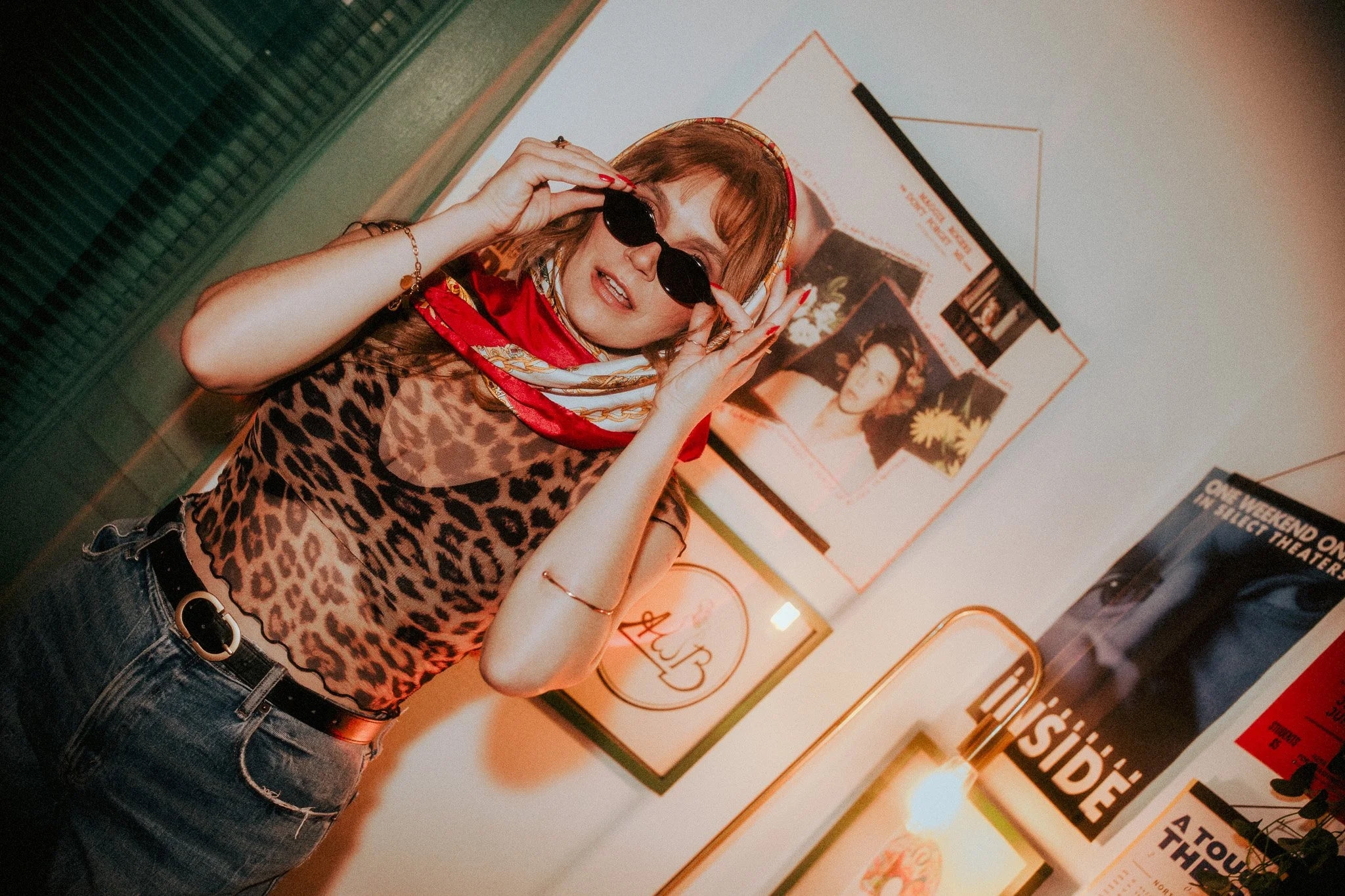 A woman wearing a leopard print top, red bandana, and sunglasses poses indoors, adjusting her sunglasses, in front of framed posters on a wall.