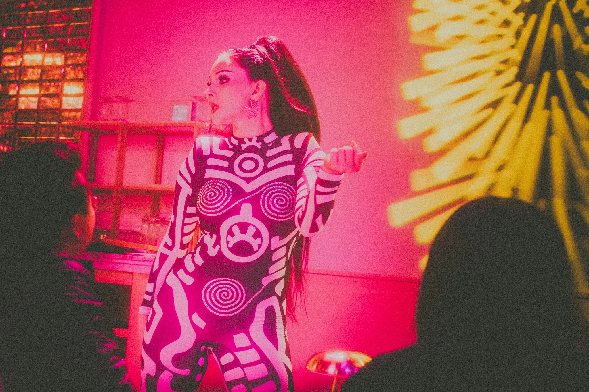 A woman with long dark hair, wearing a black and white patterned dress, stands in a pink-lit room, engaging with two seated individuals, one male and one female. The background features decorative elements and a bright yellow sunburst. The woman has 