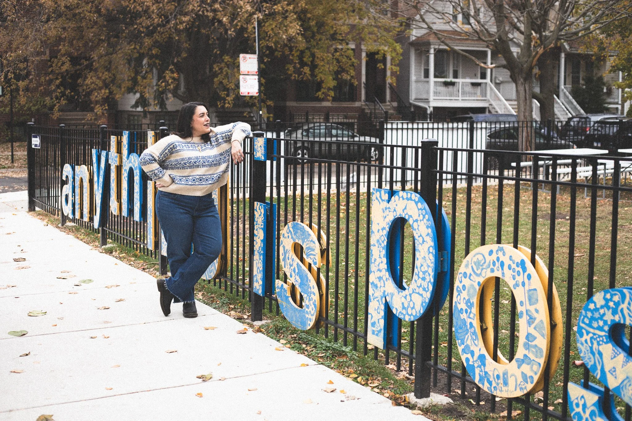 A woman in a patterned sweater and jeans leans against a black fence with large decorative letters spelling 'asm.' Trees and houses are in the background with fallen leaves on the sidewalk.