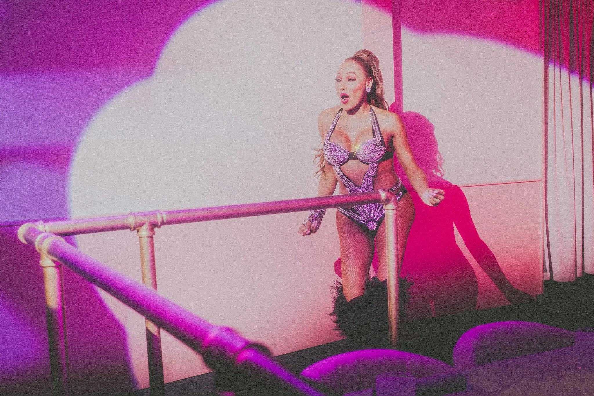 A woman in a sparkling, revealing stage costume with black fishnet tights, walking on a stage with pink and purple lighting, holding a railing and looking surprised or excited.
