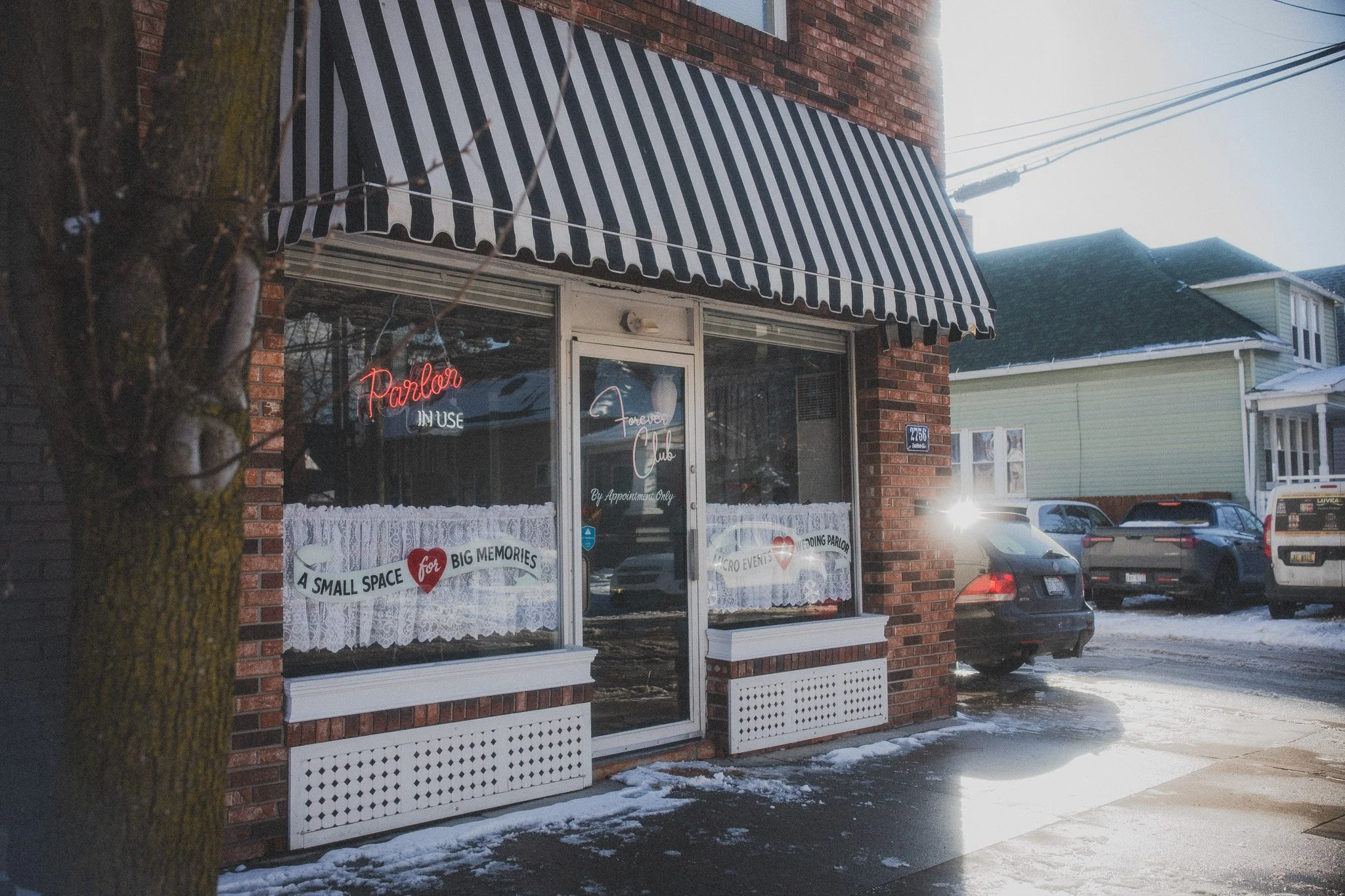A small brick storefront with a black and white striped awning. The window displays neon signs and lace curtains, with snowy pavement outside and parked cars in the background.