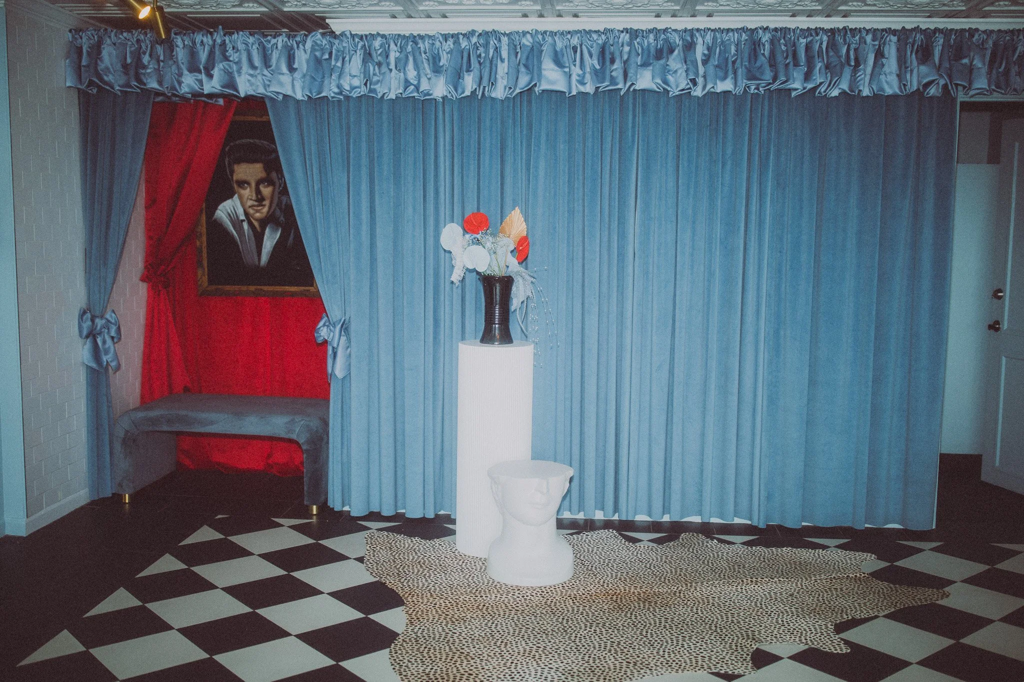 Room with blue curtains, a bench with blue upholstery, a painting of a young person on red wall, a white pedestal with a black vase containing colorful paper flowers, and a white sculpture of a head on a leopard-print rug.