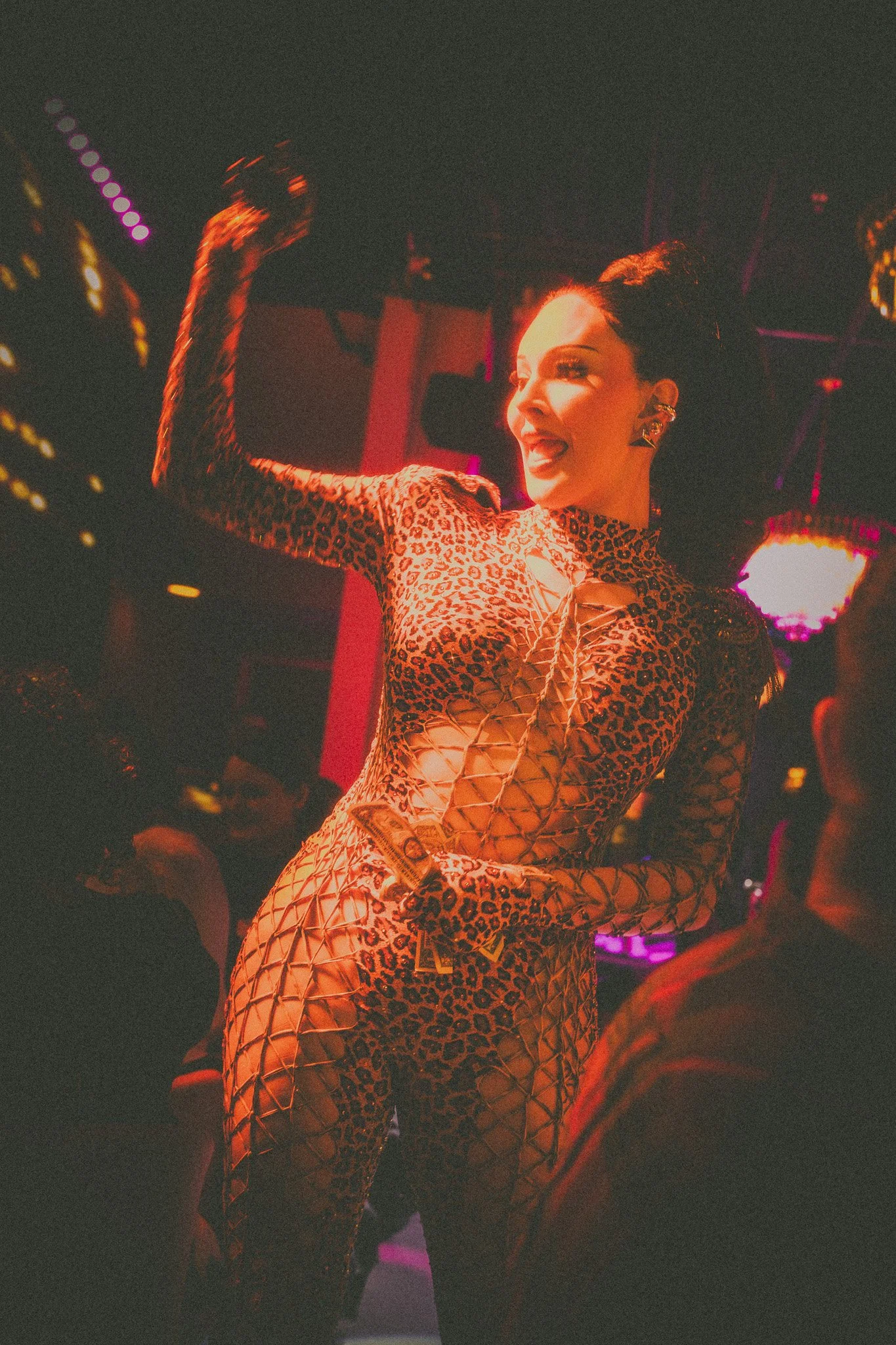 A woman in a leopard print outfit, smiling and flexing her arm in a nightclub with colorful lighting.
