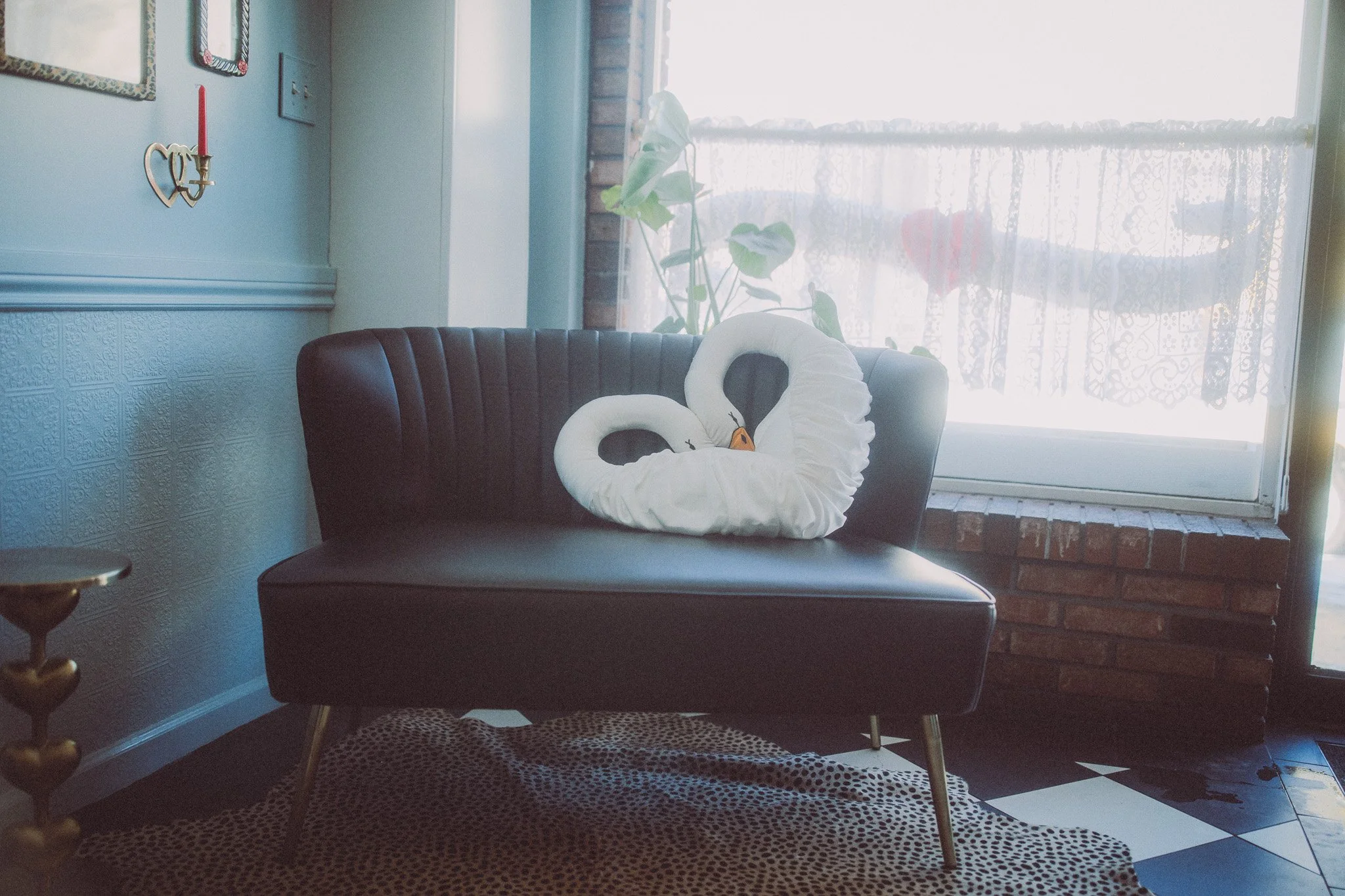 A black leather sofa with two white swan-shaped towel art pieces on it, positioned near a window with lace curtains, a small potted plant, and a brick wall.