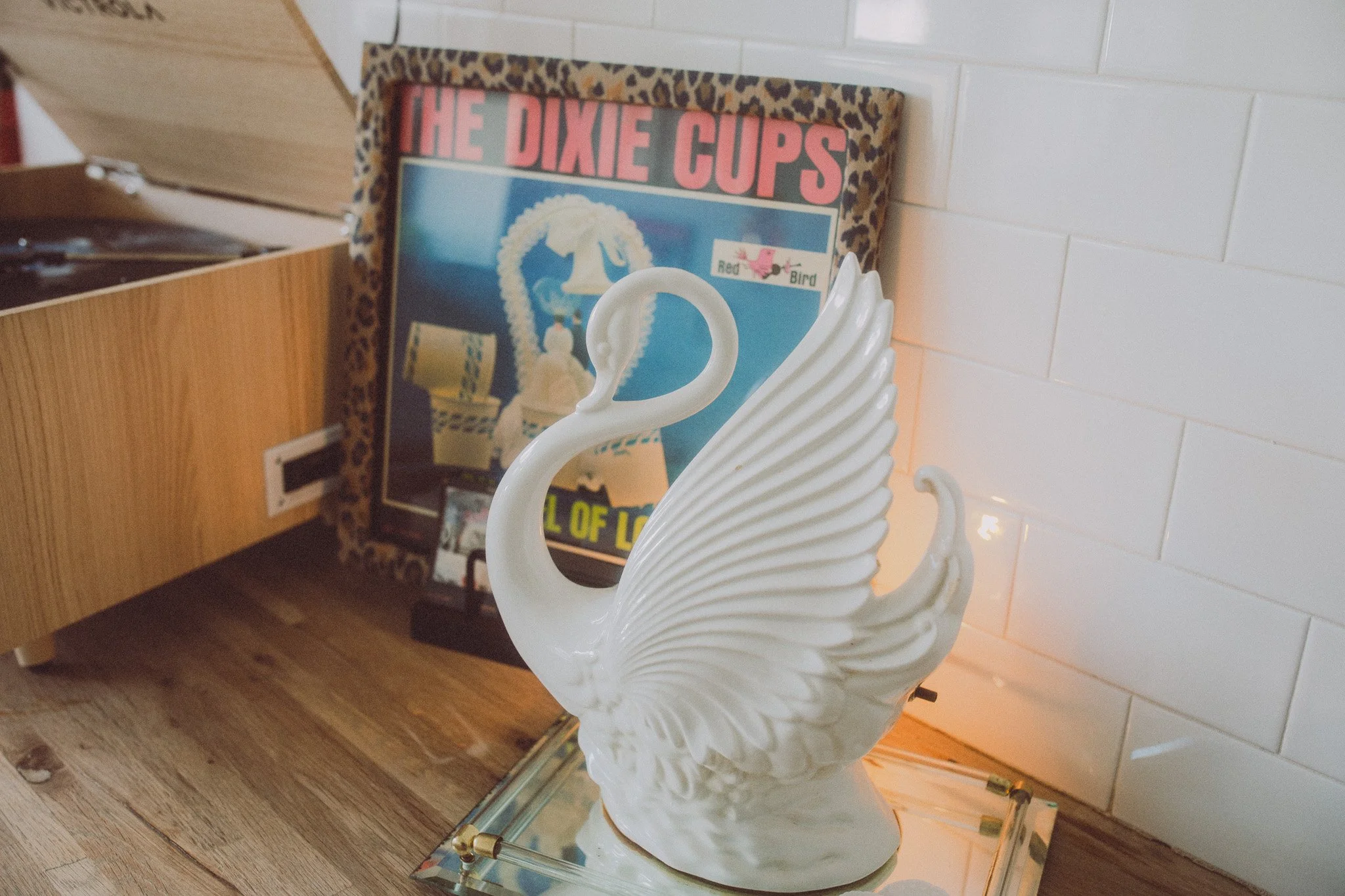 A white swan-shaped decorative figurine with detailed feather wings and neck, placed on a glass surface in front of a framed poster that reads "The Dixie Cups" and features a phone with a rotary dial, a cassette tape, and a pink bird sticker, set aga
