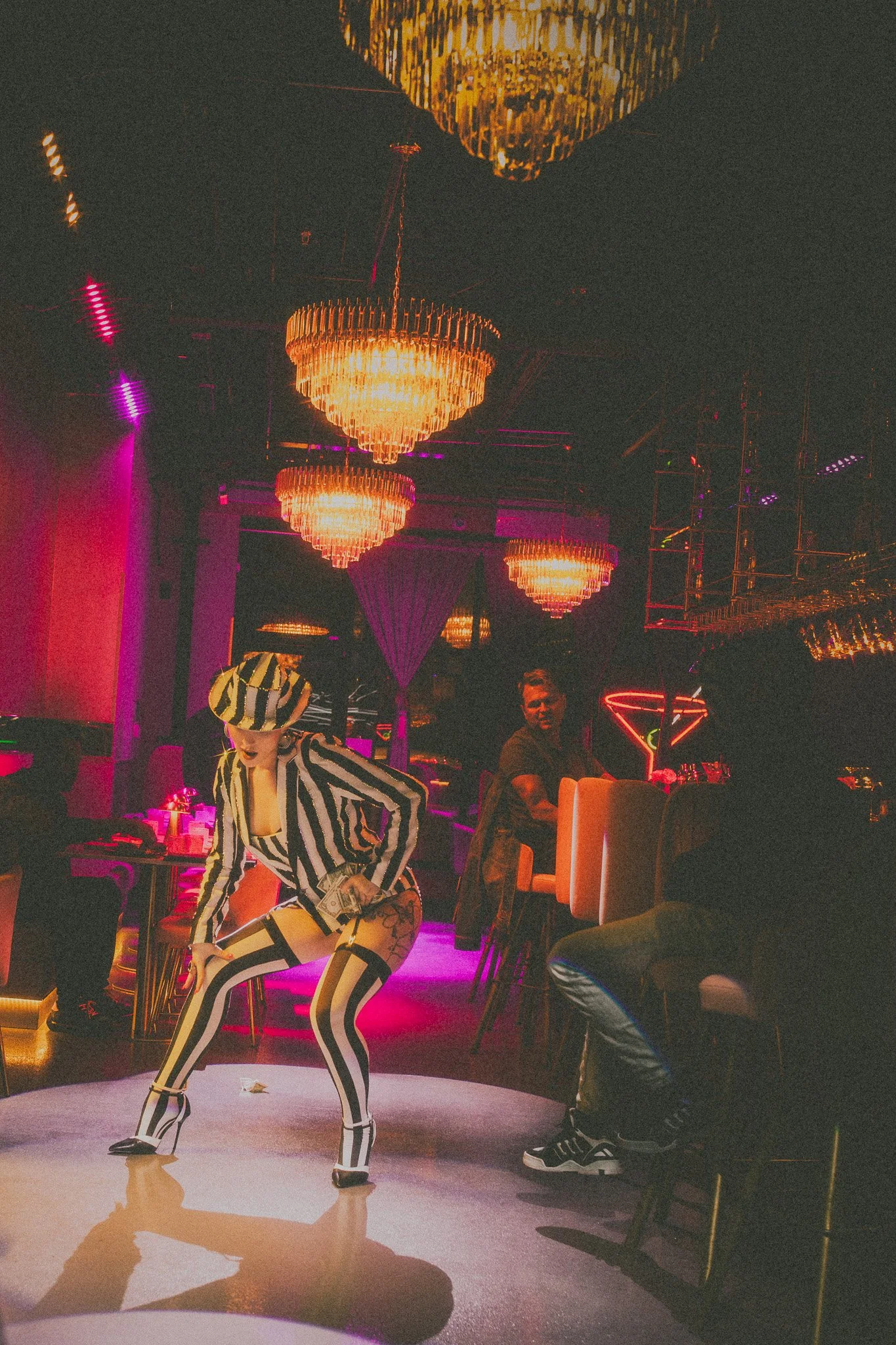 A person dancing at a nightclub with pink and orange lighting, chandeliers on the ceiling, and people sitting at tables in the background.