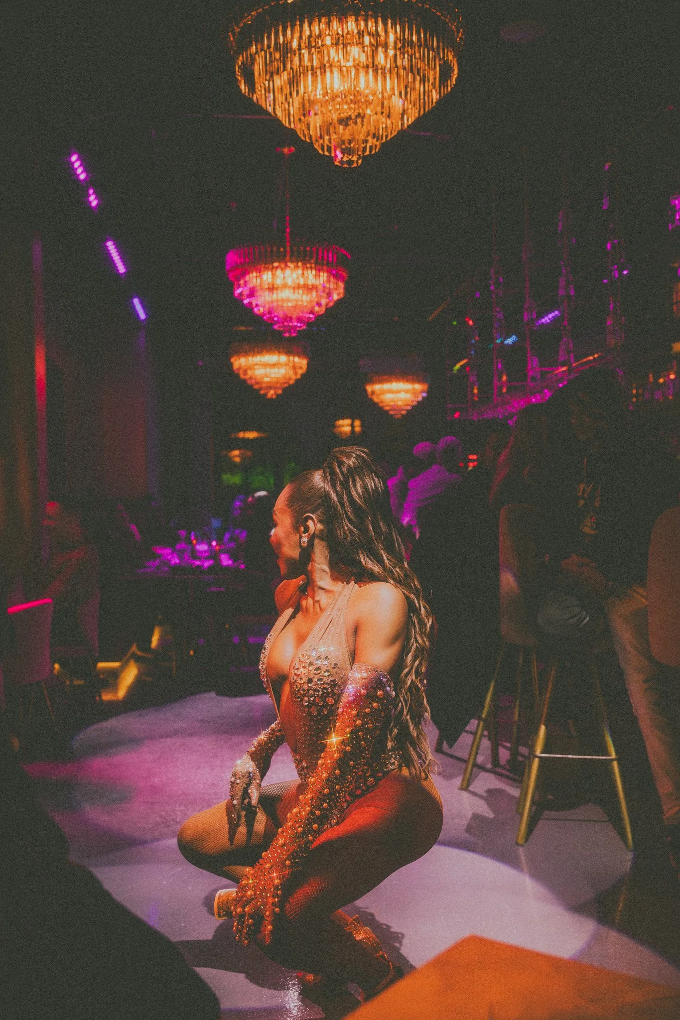 A woman with long hair in a sparkly, embellished gown with sheer sleeves is squatting on a stage in a dimly lit club or event venue with chandeliers and people in the background.