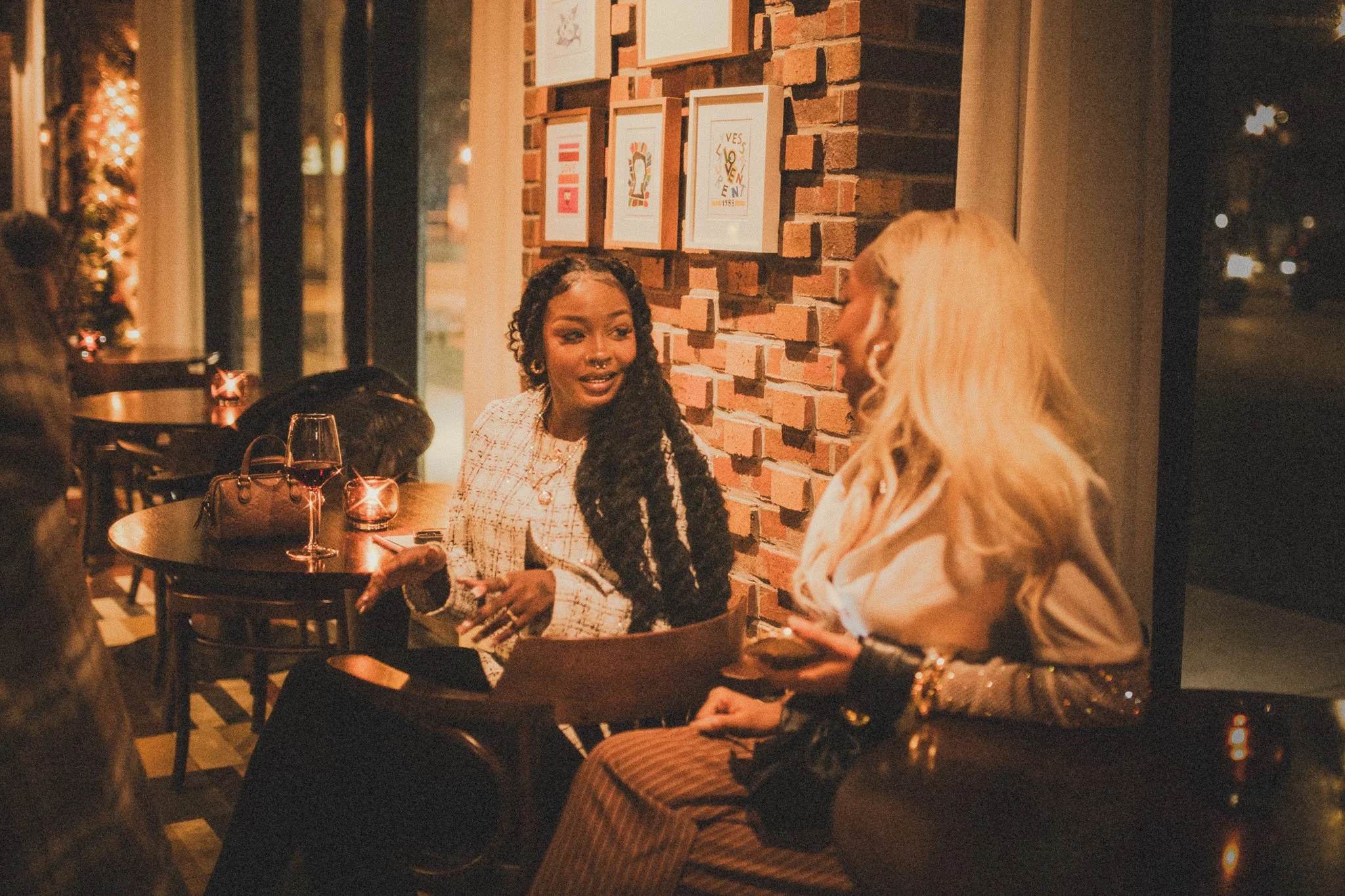 Two women having a conversation at a restaurant table, with a brick wall decorated with framed artwork in the background, and a glass of red wine on the table.