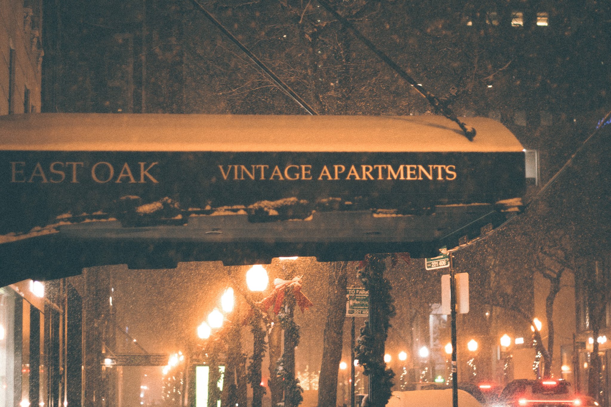 Street scene at night with illuminated streetlights, parked cars, and trees decorated for the holidays, seen through a rain-covered window. A sign reads "East Oak Vintage Apartments."