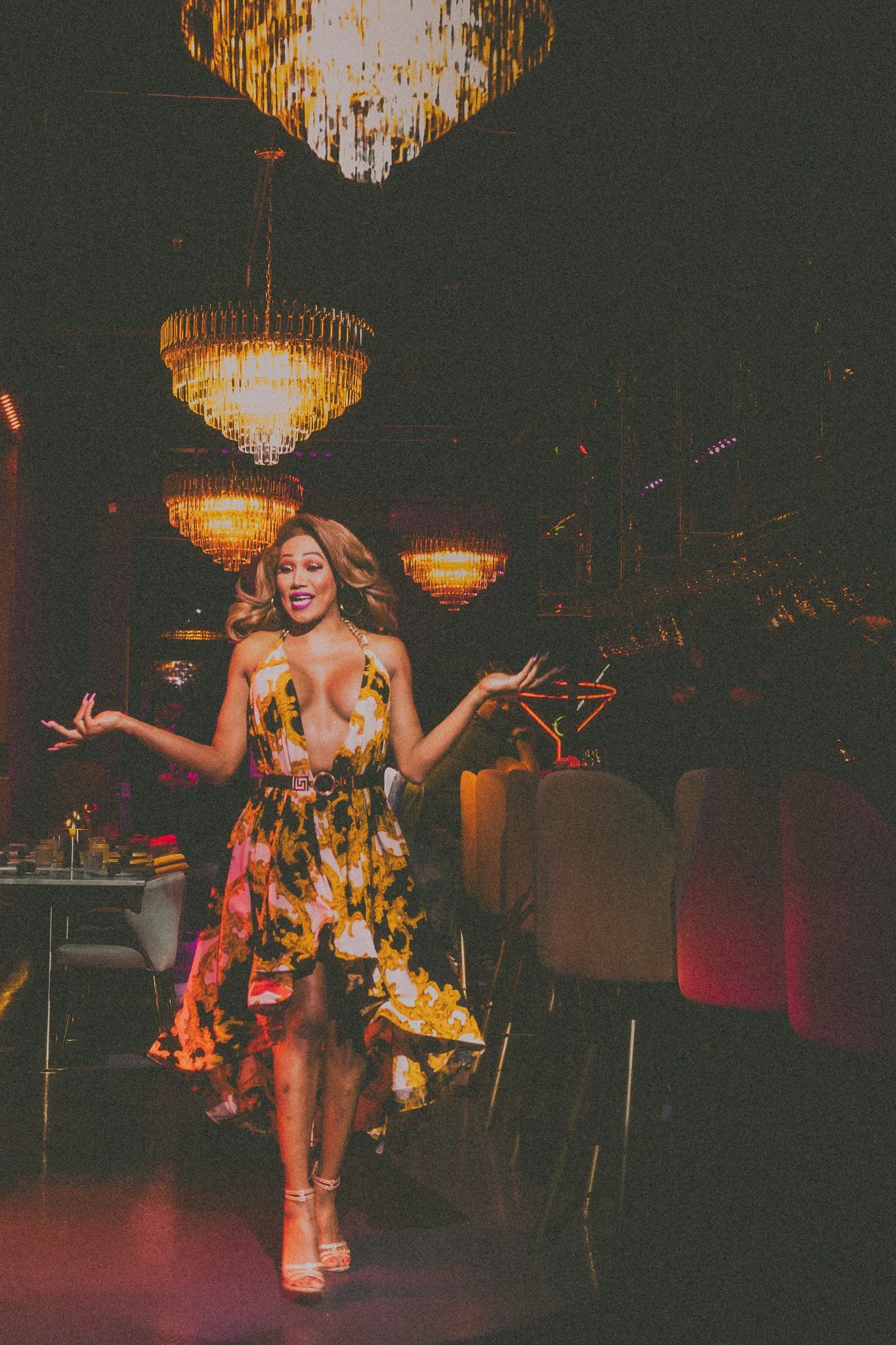 A woman dancing in a vibrant yellow and black patterned dress in a dimly lit nightclub with chandeliers and purple accent lighting.