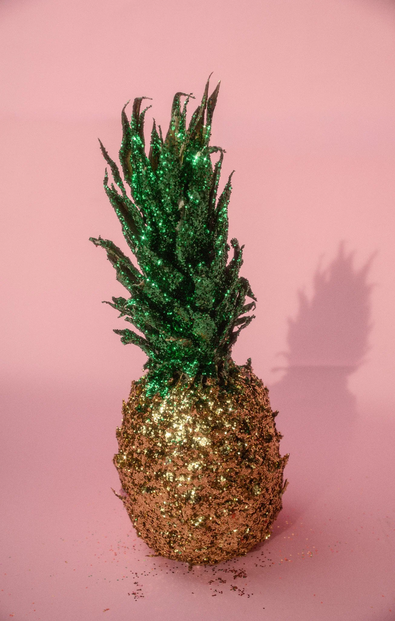 Decorative pineapple covered in gold glitter, with green glittery leaves, set against a pink background, casting a shadow.