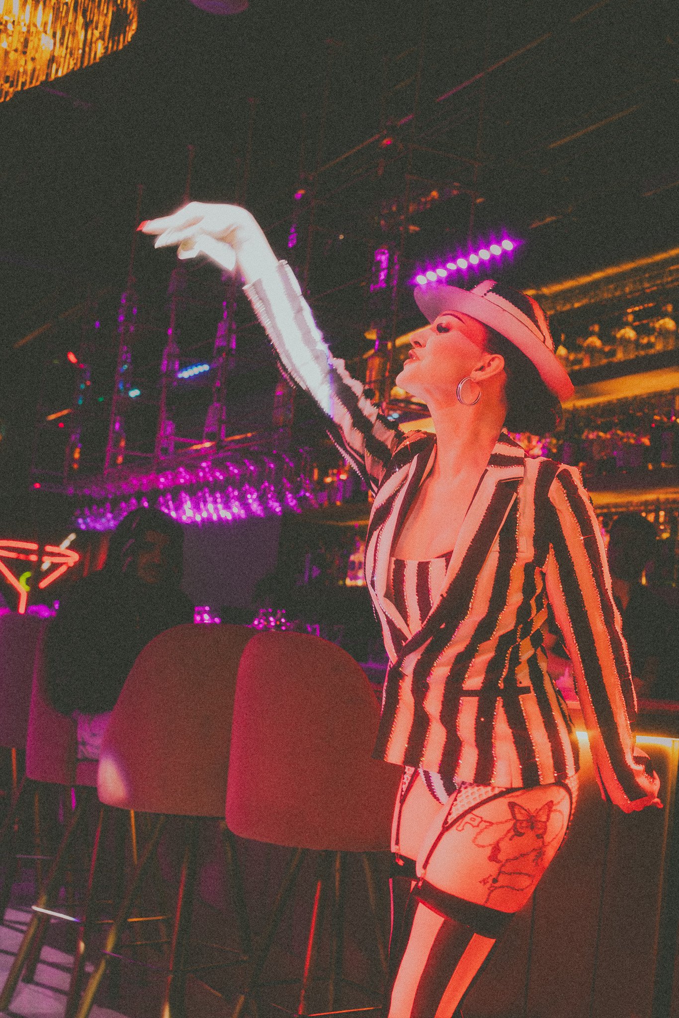 A woman in a striped blazer and matching shorts with thigh-high stockings and a tattoo, dancing at a colorful nightclub with neon lights and a DJ in the background.