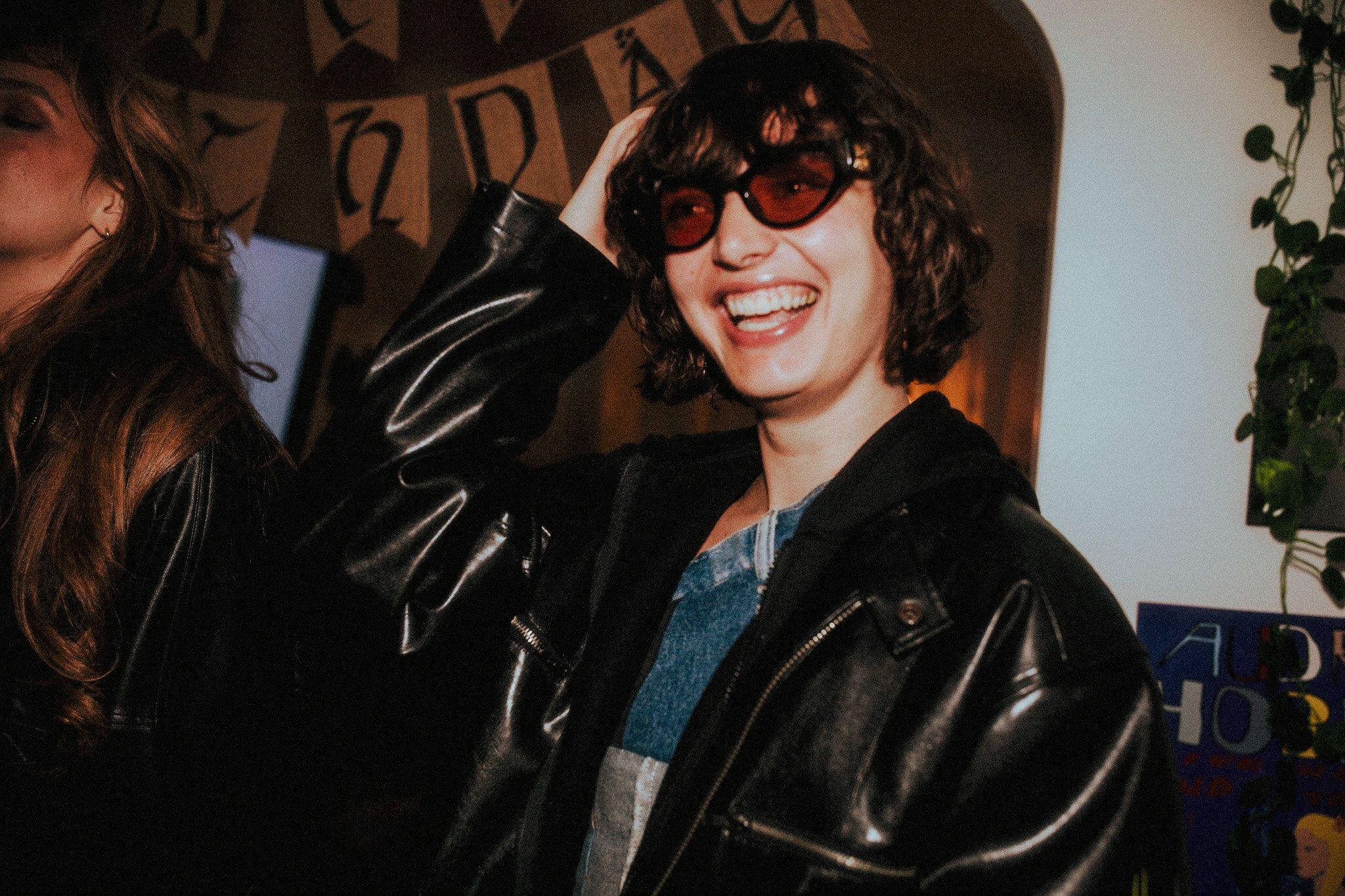 A woman with curly dark hair wearing sunglasses, a black leather jacket, and a blue denim shirt, smiling and touching her head in a social setting.