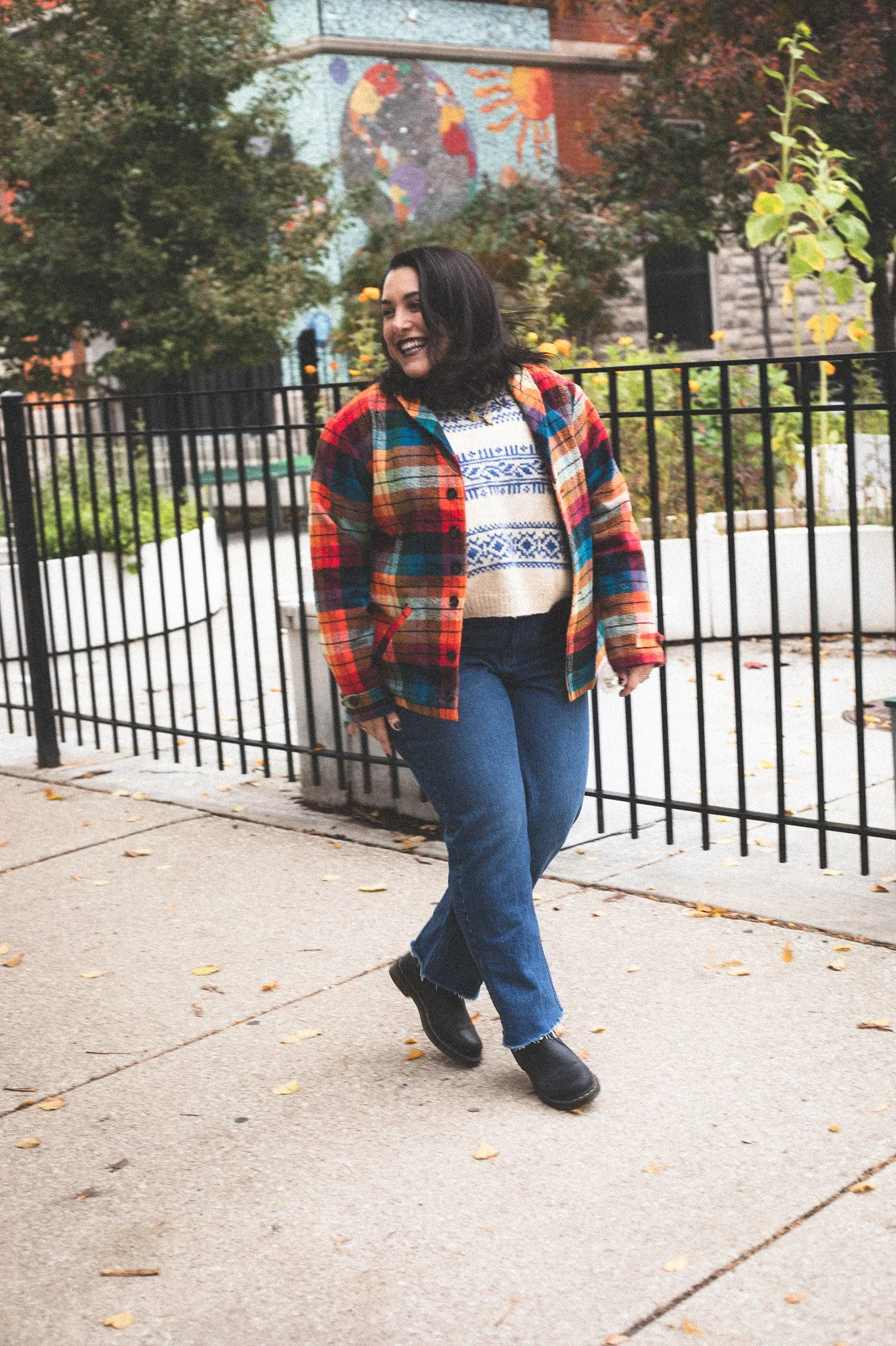 A woman smiling and walking outdoors on a city sidewalk, wearing a colorful plaid jacket, a patterned sweater, jeans, and black boots, with trees, a fence, and a mural in the background.